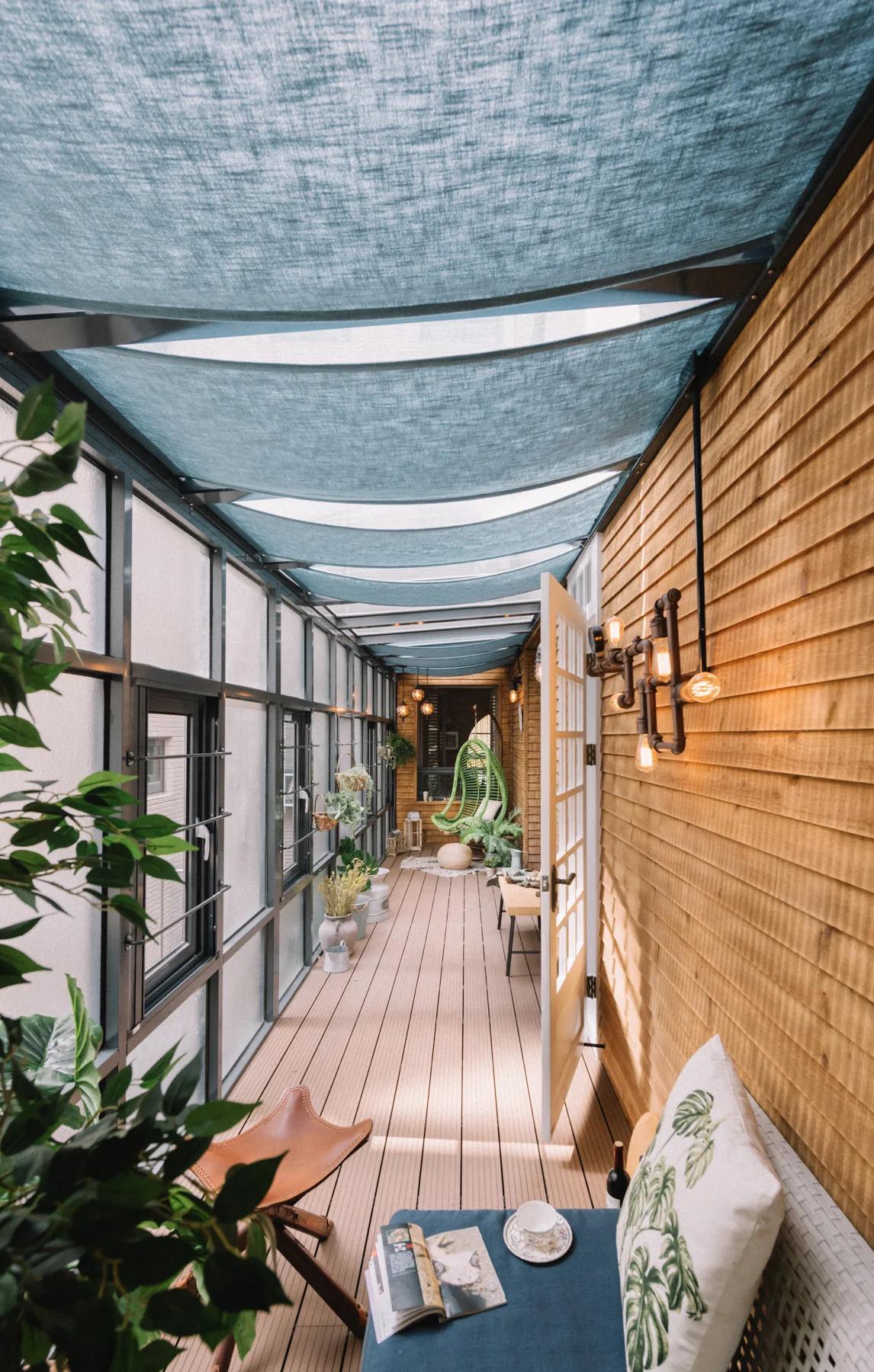 A long enclosed porch features wood-paneled walls, a blue fabric canopy overhead, a glass wall, and outdoor seating including a hanging chair.