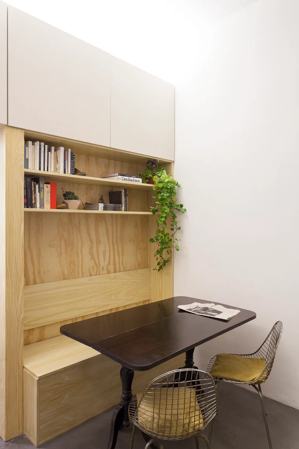 A compact dining area featuring a light wood built-in storage unit with bench seating, a dark table, and two metal wire chairs.
