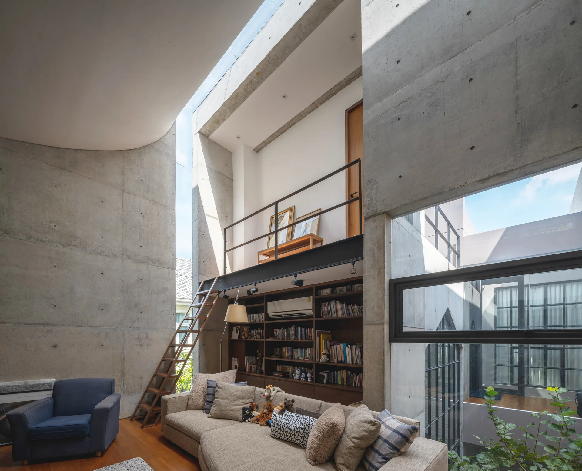 A double-height living room featuring exposed concrete walls, a large wooden bookcase beneath a mezzanine level, a sectional sofa, and a skylight.