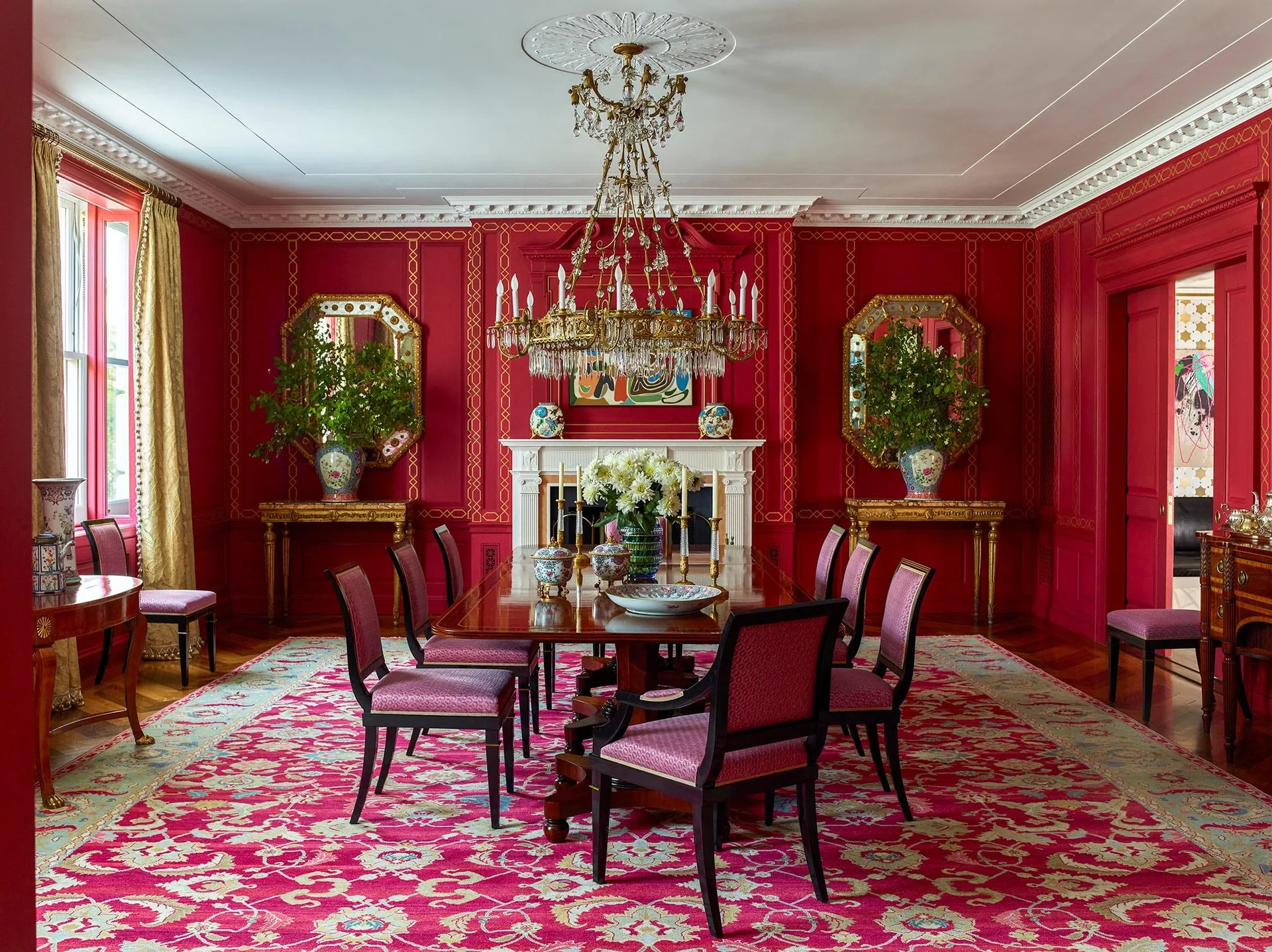 A formal dining room features vibrant red walls with gold trim, a long wood table under a crystal chandelier, and a pink patterned rug.