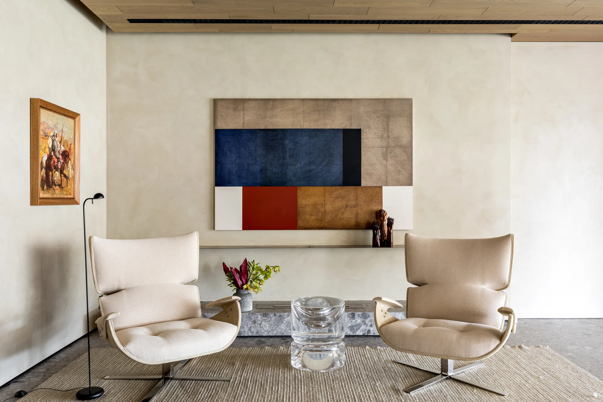 Two cream upholstered swivel armchairs face a clear acrylic side table on a textured beige rug in front of a large geometric painting.