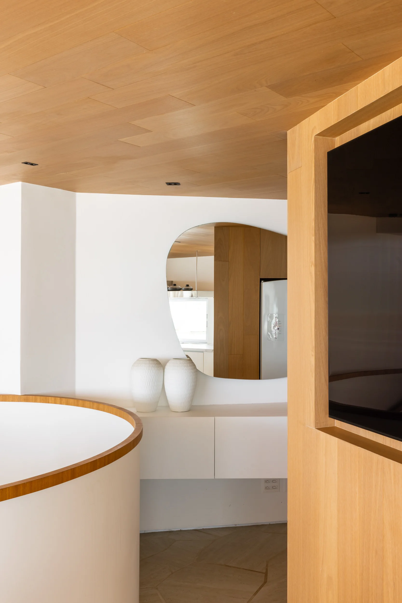 A modern interior space features a light wood-paneled ceiling, a curved white partition wall, a floating cabinet, and an irregular mirror.