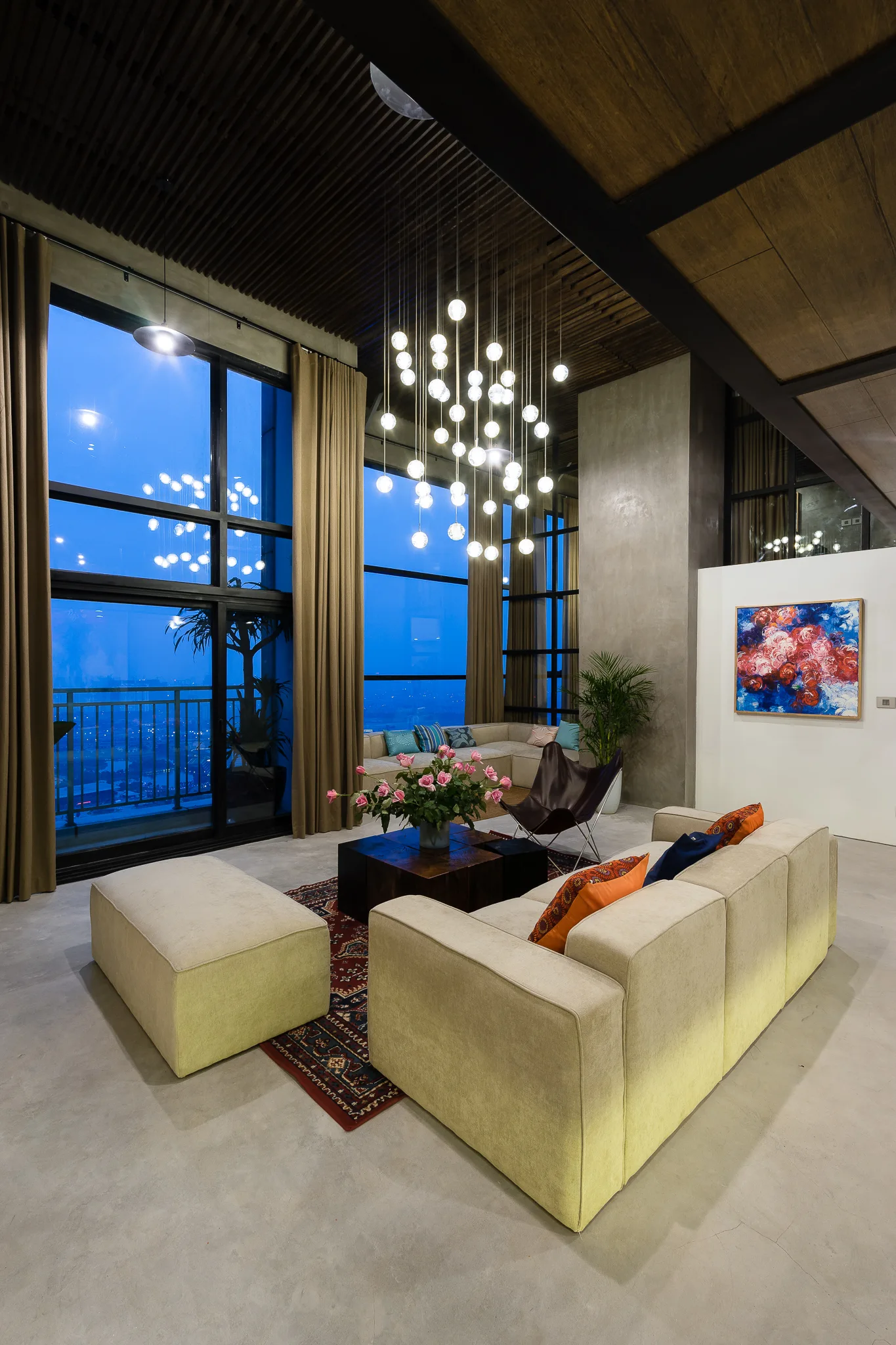 A modern double-height living room featuring concrete floors, beige modular sofas, a large multi-globe chandelier, and tall windows.