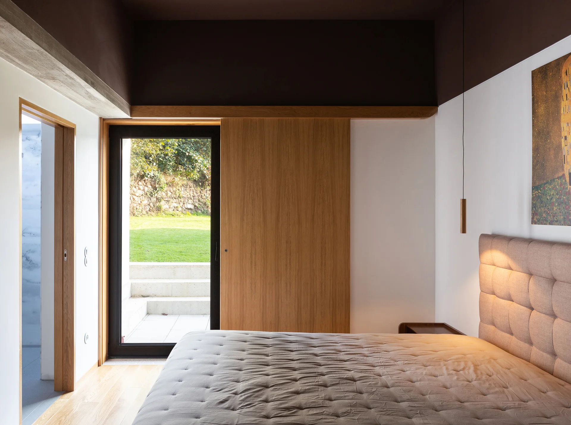 A modern bedroom featuring a beige tufted headboard, light wood floors, and a dark brown painted ceiling with a glass door opening to a garden.