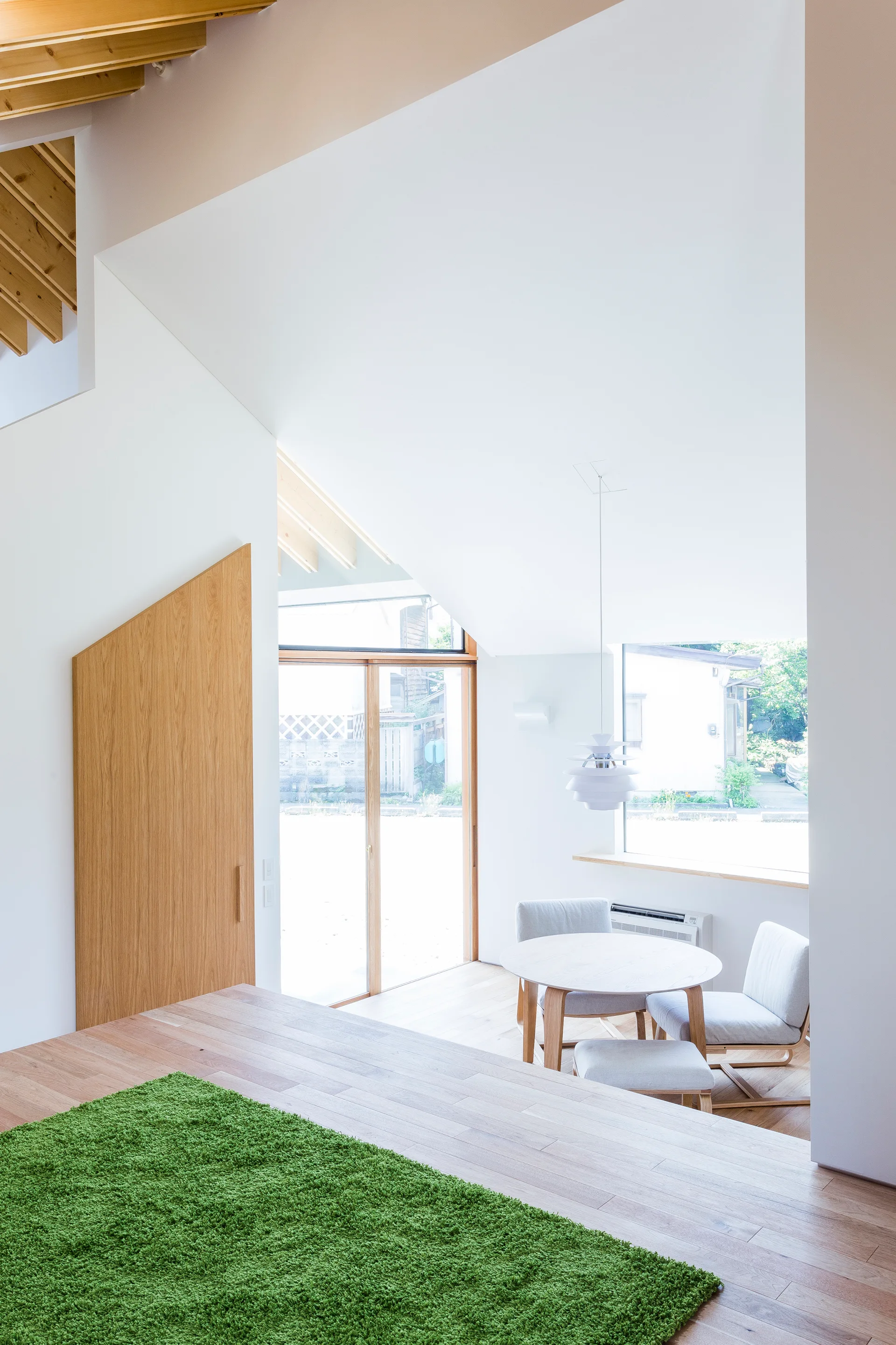 A light-filled dining room features a round wooden table, upholstered chairs under a sloped ceiling, and a bright green rug on the wood floor.