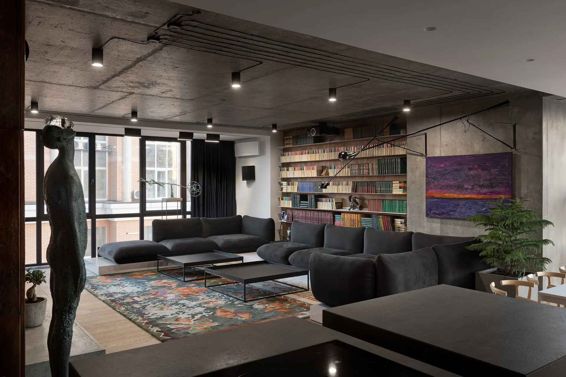 A modern industrial living room features a dark charcoal sectional sofa, an exposed concrete ceiling with track lighting, and a large built-in bookshelf.