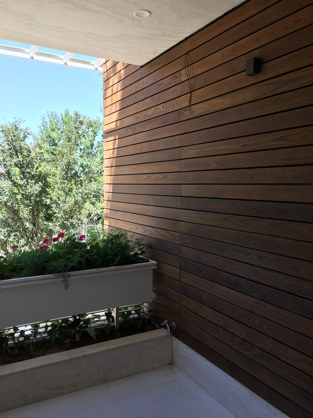 A modern balcony features a horizontal wood slat wall, a white planter box filled with green foliage and pink flowers, and a light grey concrete ceiling.
