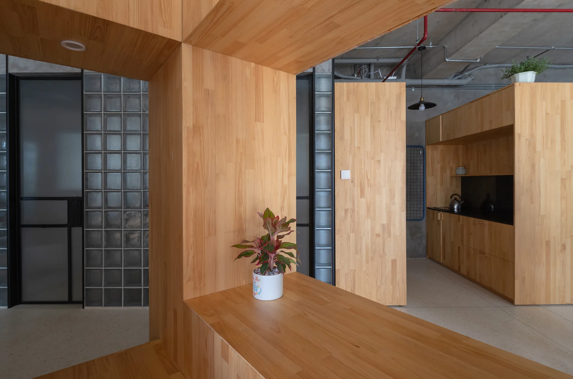 A minimalist kitchen featuring custom wood cabinetry, a large matching wood partition, a glass block wall, and an exposed concrete ceiling with pipes.