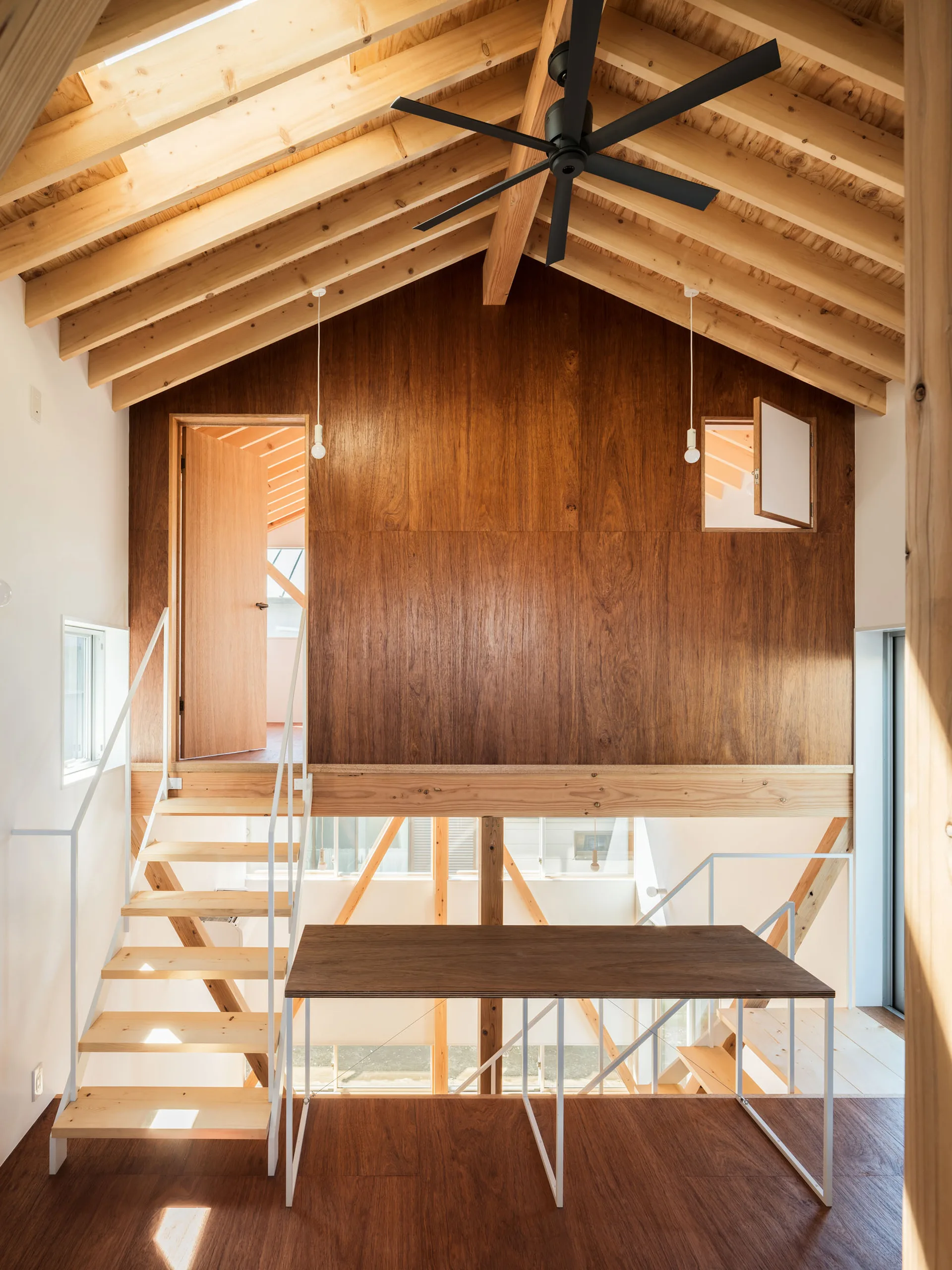 A minimalist loft interior features exposed light wood rafters, a central dark wood paneled wall with a doorway, and a matching wooden staircase.
