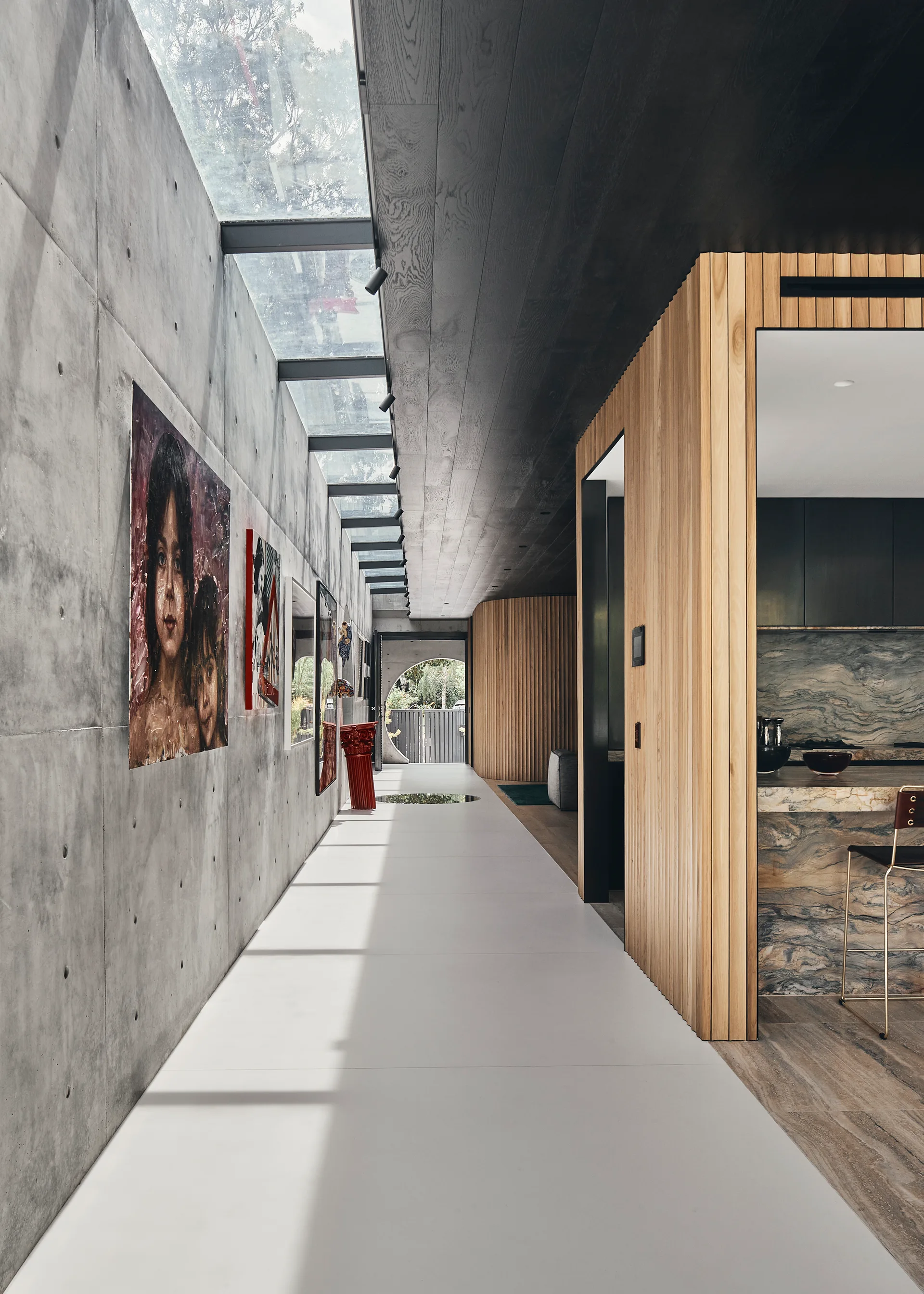 A long modern hallway featuring an exposed concrete wall with artwork, a continuous glass skylight, and vertical wood slat paneling.