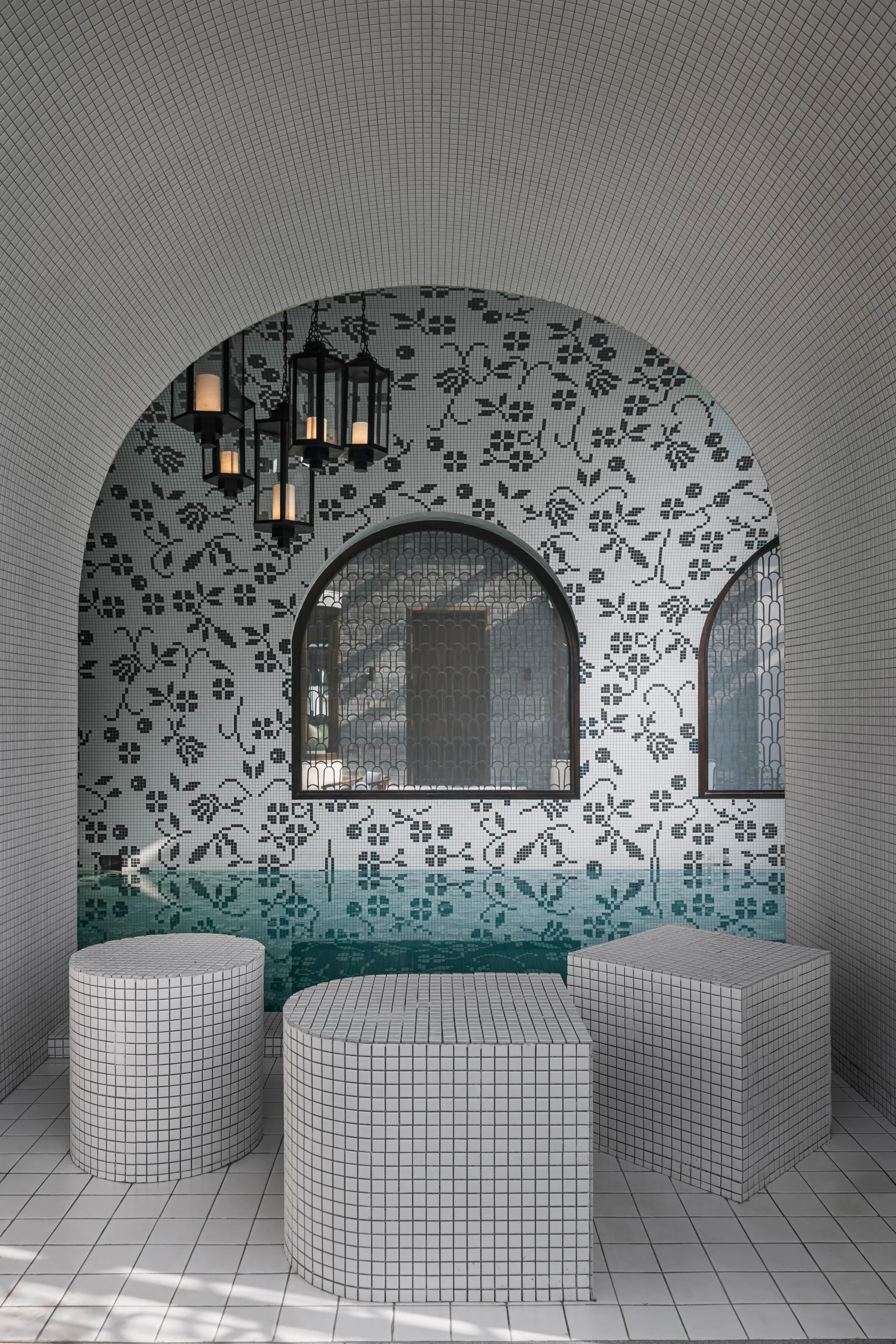 An indoor pool area with white tiled arched walls, a black floral mosaic pattern, hanging lanterns, and three geometric tile-clad stools.