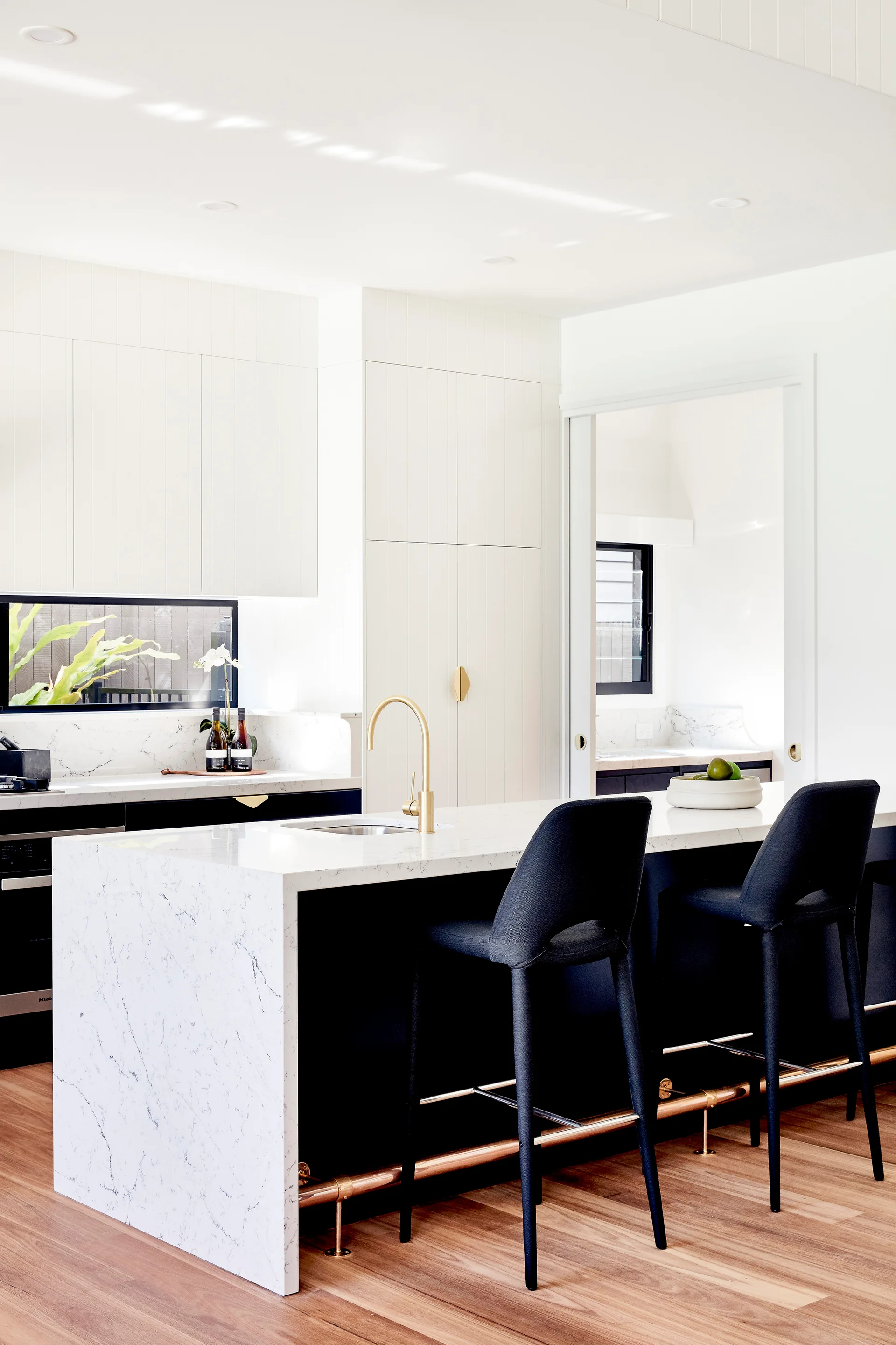A modern kitchen features a marble waterfall island with black stools, a gold faucet, a brass footrail, and integrated white cabinetry.