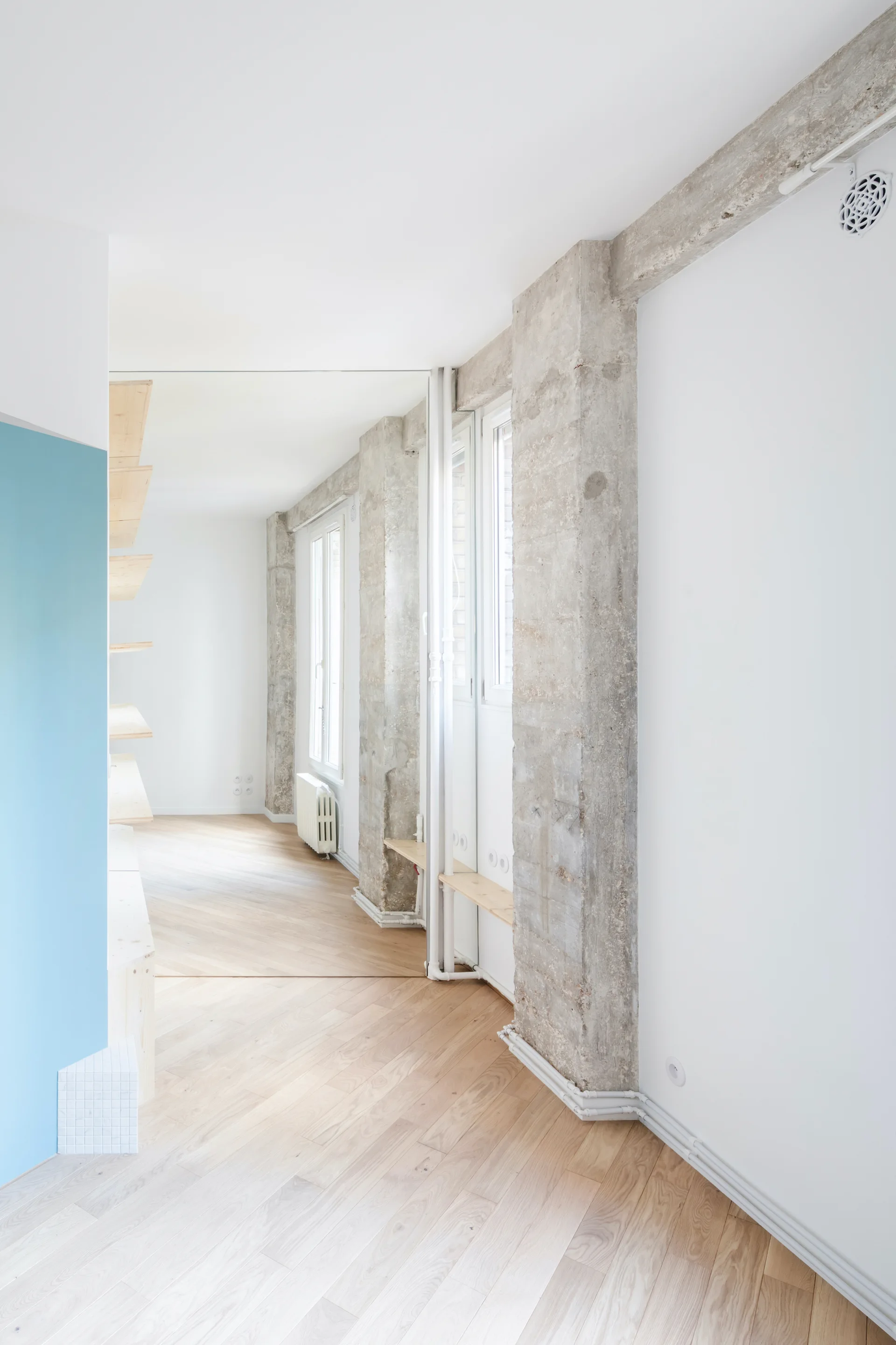 A minimalist room with light wood floors, stark white walls, raw concrete pillars, and a floor-to-ceiling mirror.