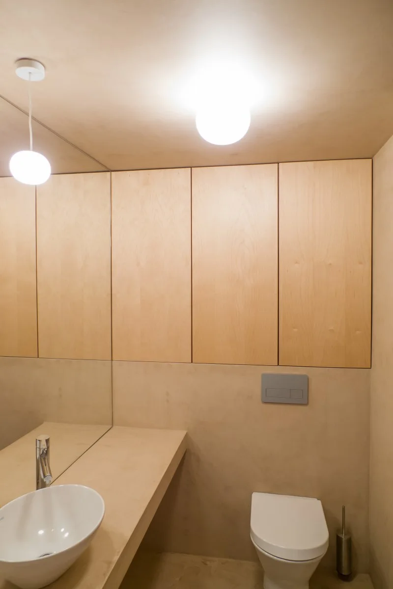 A minimalist powder room features a white wall-mounted toilet, an integrated beige vanity with a white vessel sink, and flush-mounted light wood cabinets.