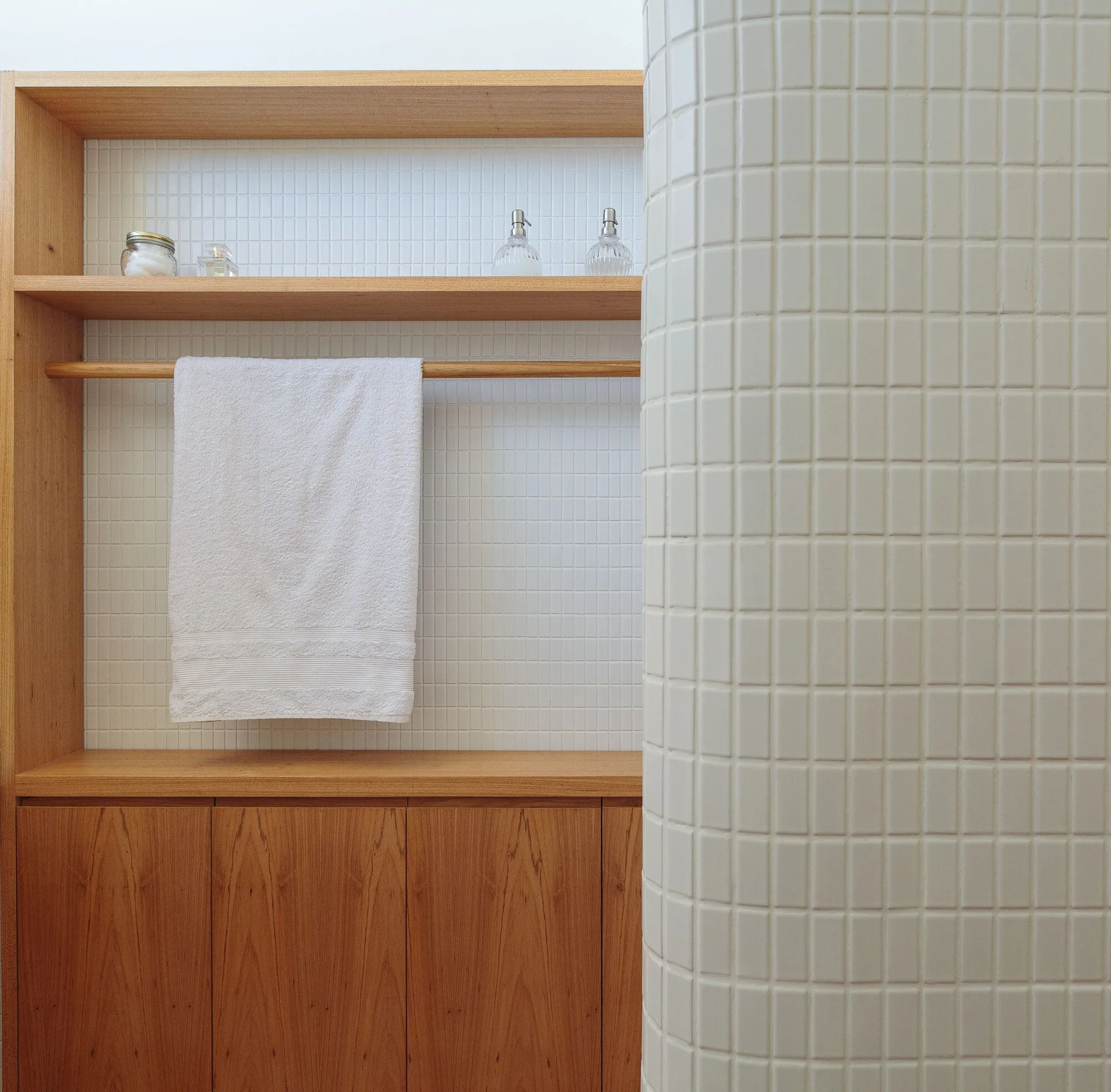 A bathroom view of a built-in wood cabinet with open shelves and a towel rail, next to a curved partition wall in white grid tile.