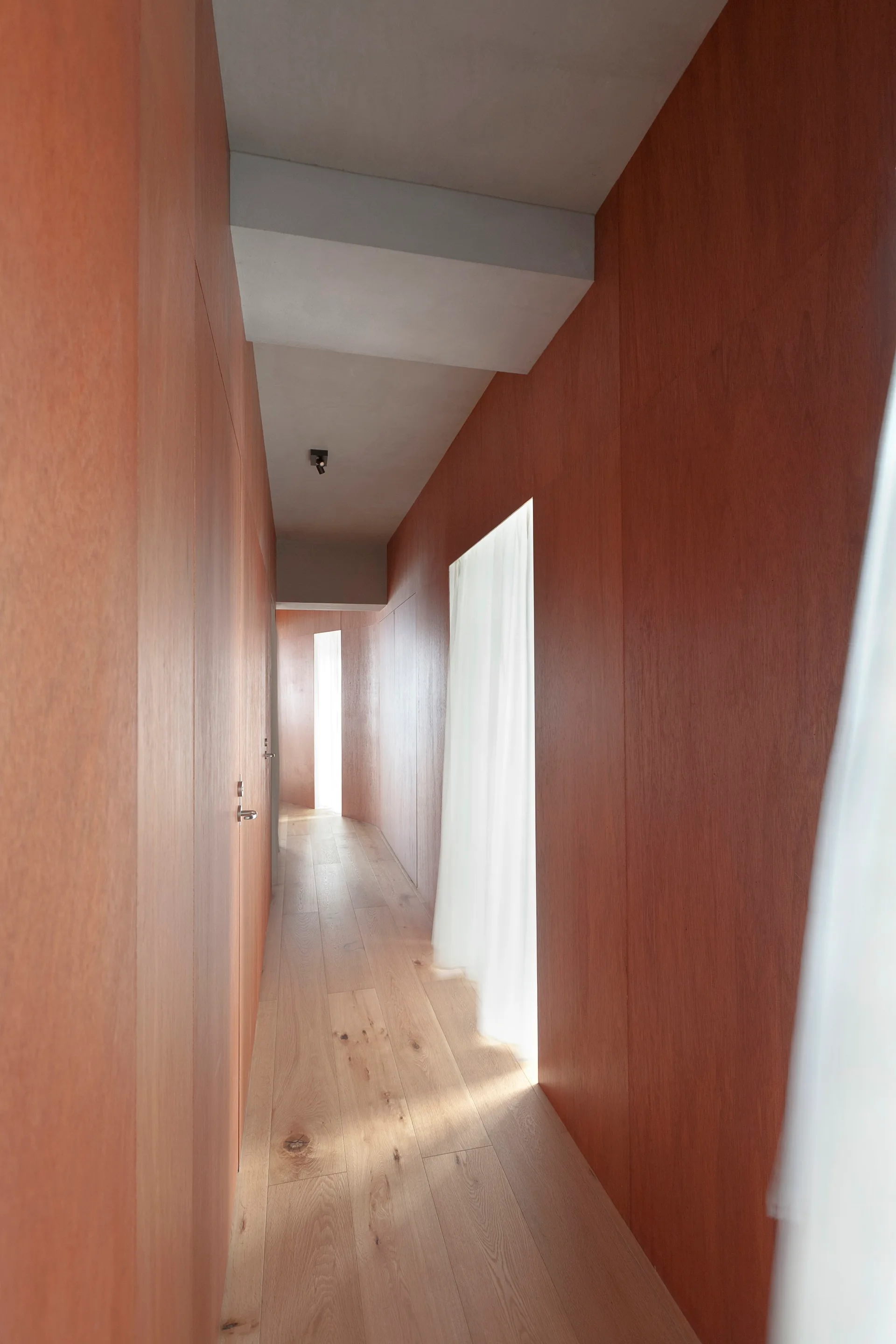 A long hallway with curved wood-paneled walls, light wood plank floors, and sheer white curtains hanging in recessed doorway openings.