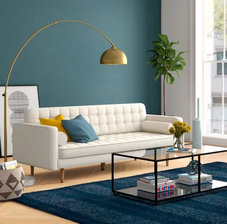 A cream tufted sofa sits on a deep blue rug against a dark teal wall, highlighted by a large arched brass floor lamp.