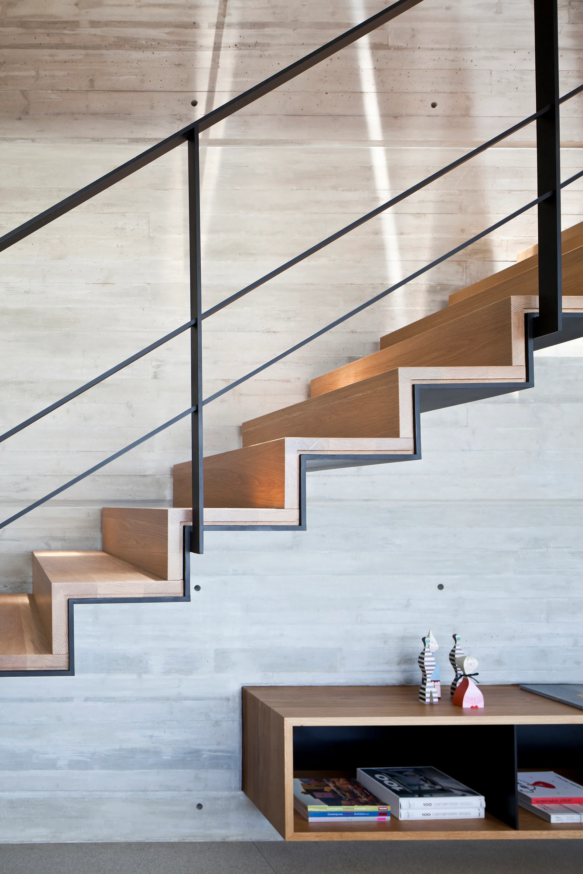 A modern wooden staircase with a thin black metal railing rises against a light grey textured concrete wall above a low wooden console table.