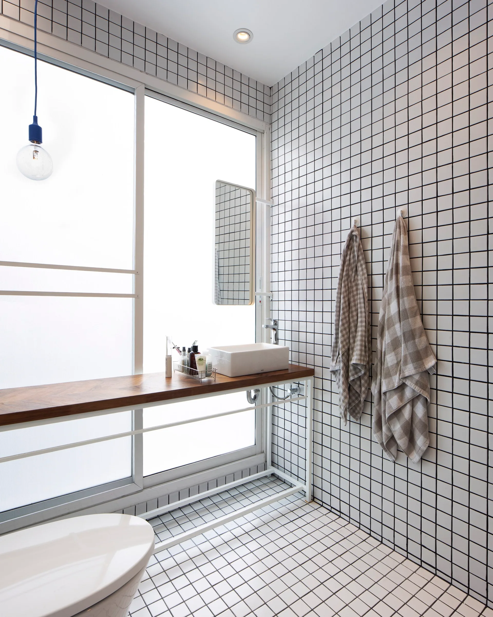 A minimalist bathroom with white square wall and floor tiles, a slender wood vanity counter, and a large frosted sliding window.