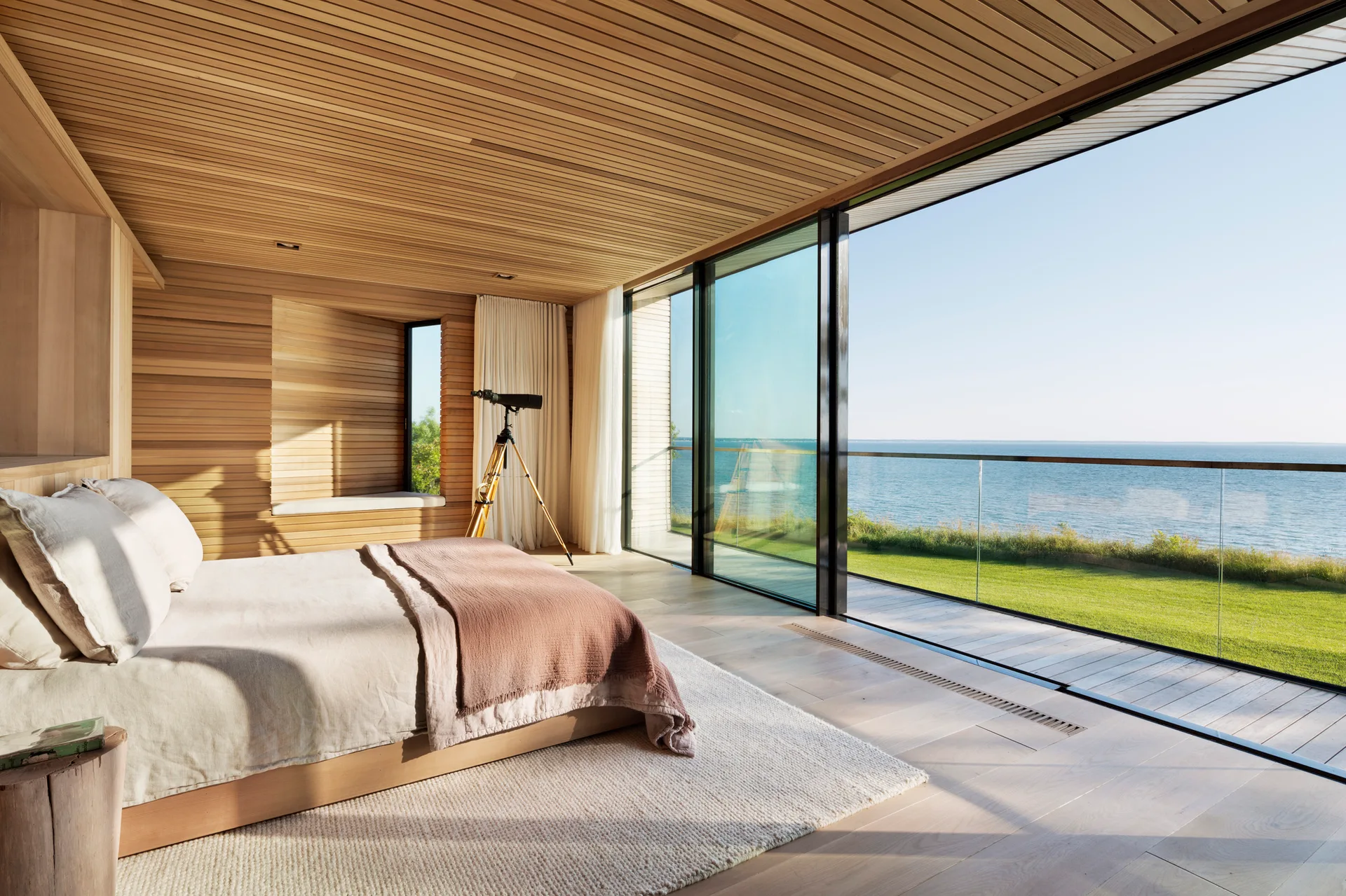 Wood-paneled bedroom featuring a low-profile bed and large floor-to-ceiling glass sliding doors that reveal a grassy lawn and ocean view.