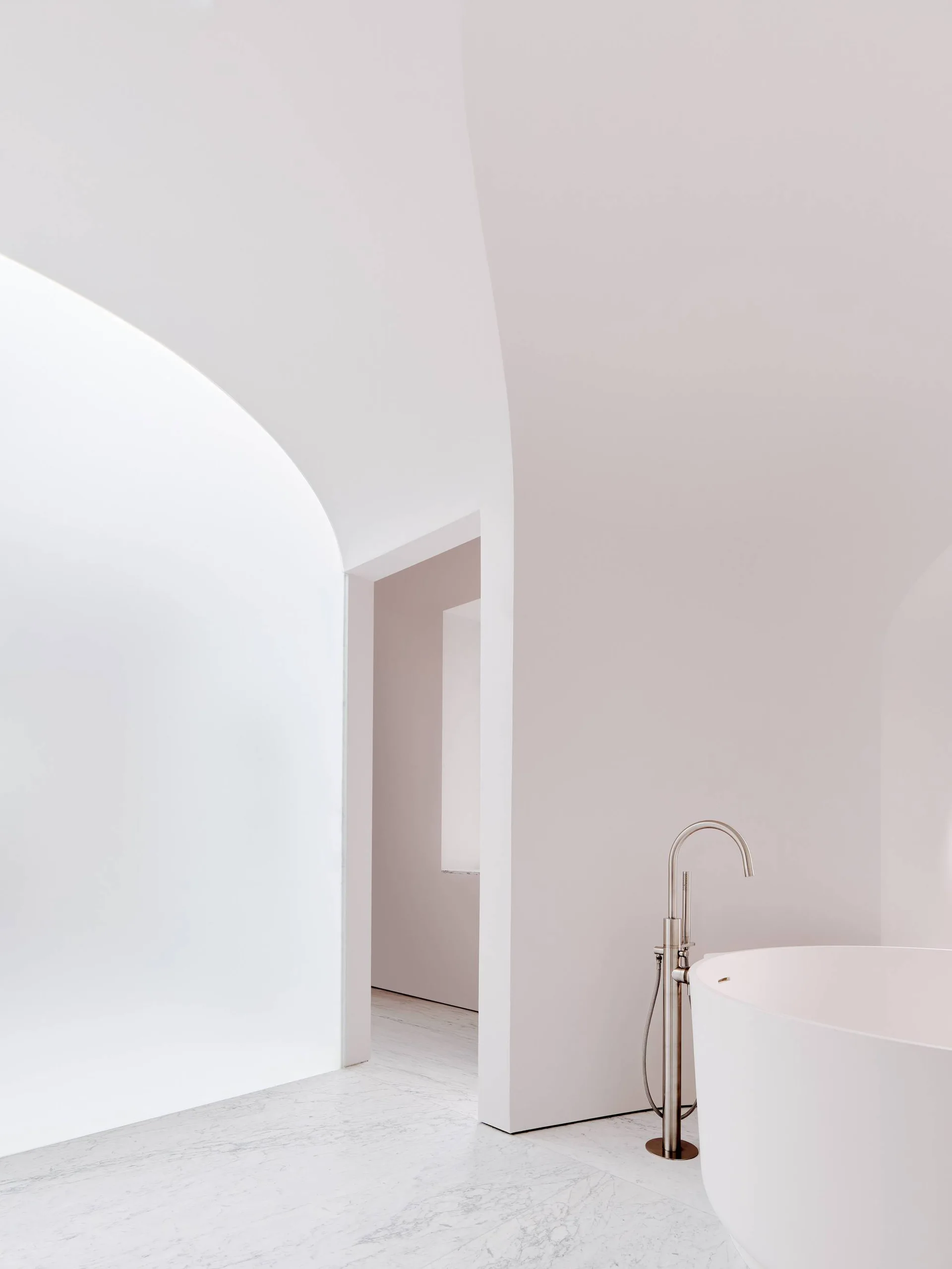 A minimalist white bathroom features a freestanding bathtub, a sleek metal faucet, and polished marble floors beneath a sweeping plaster archway.