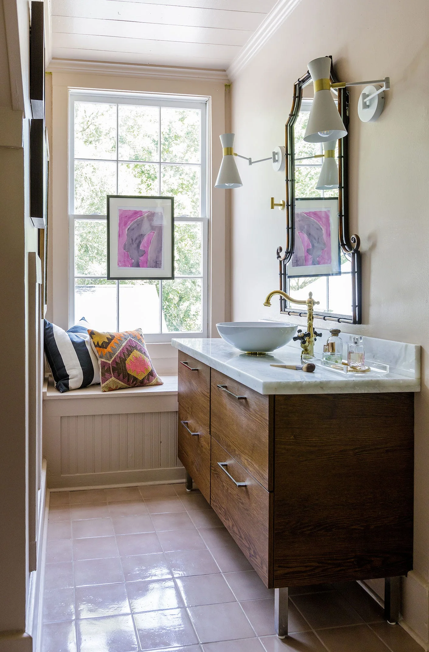 A bathroom features a dark wood vanity with a white vessel sink, brass faucet, a framed mirror, and a built-in window bench with colorful pillows.