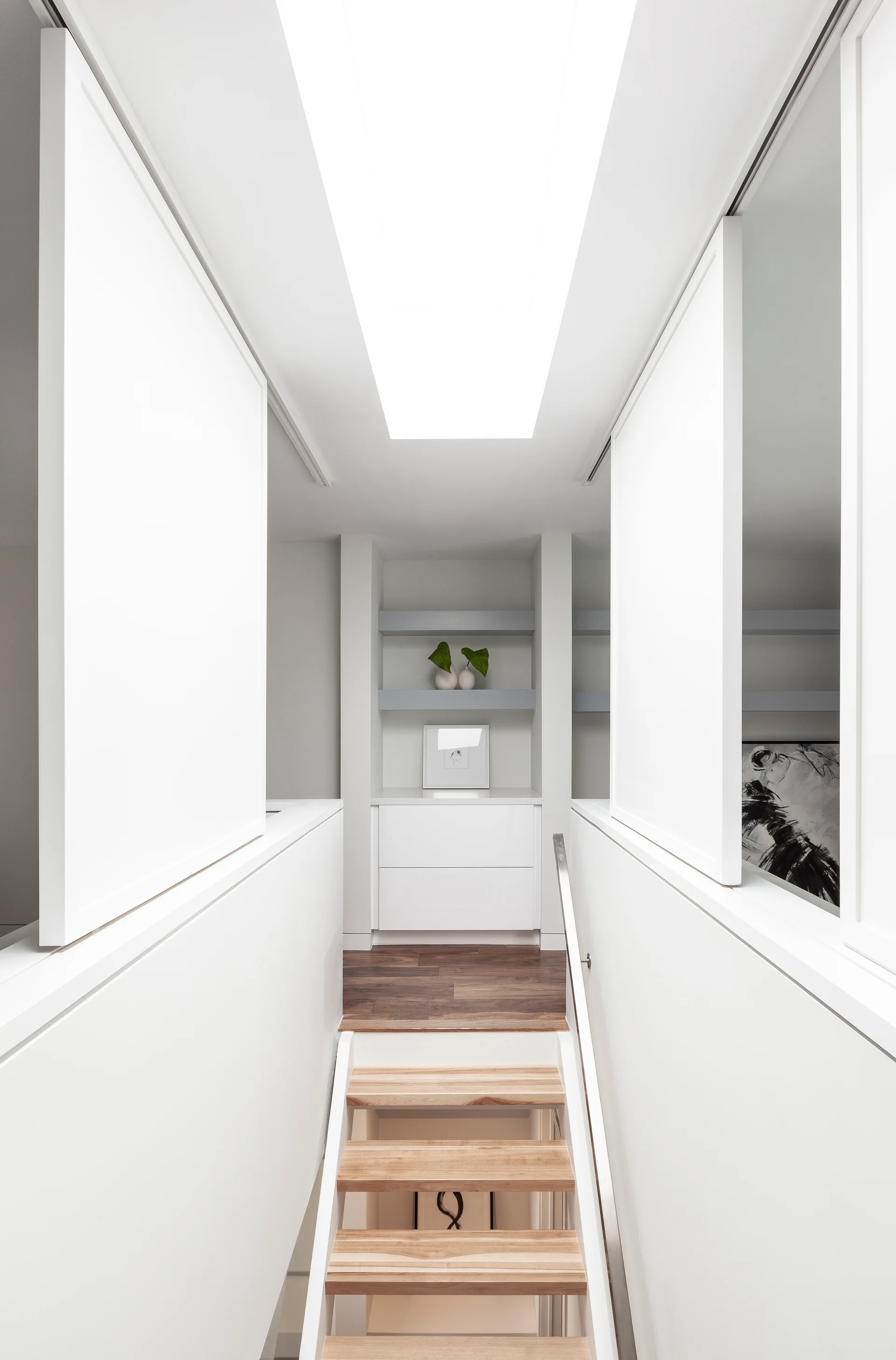 A bright white staircase landing features an open-riser wooden staircase, sliding wall panels, built-in storage, and a large rectangular skylight.