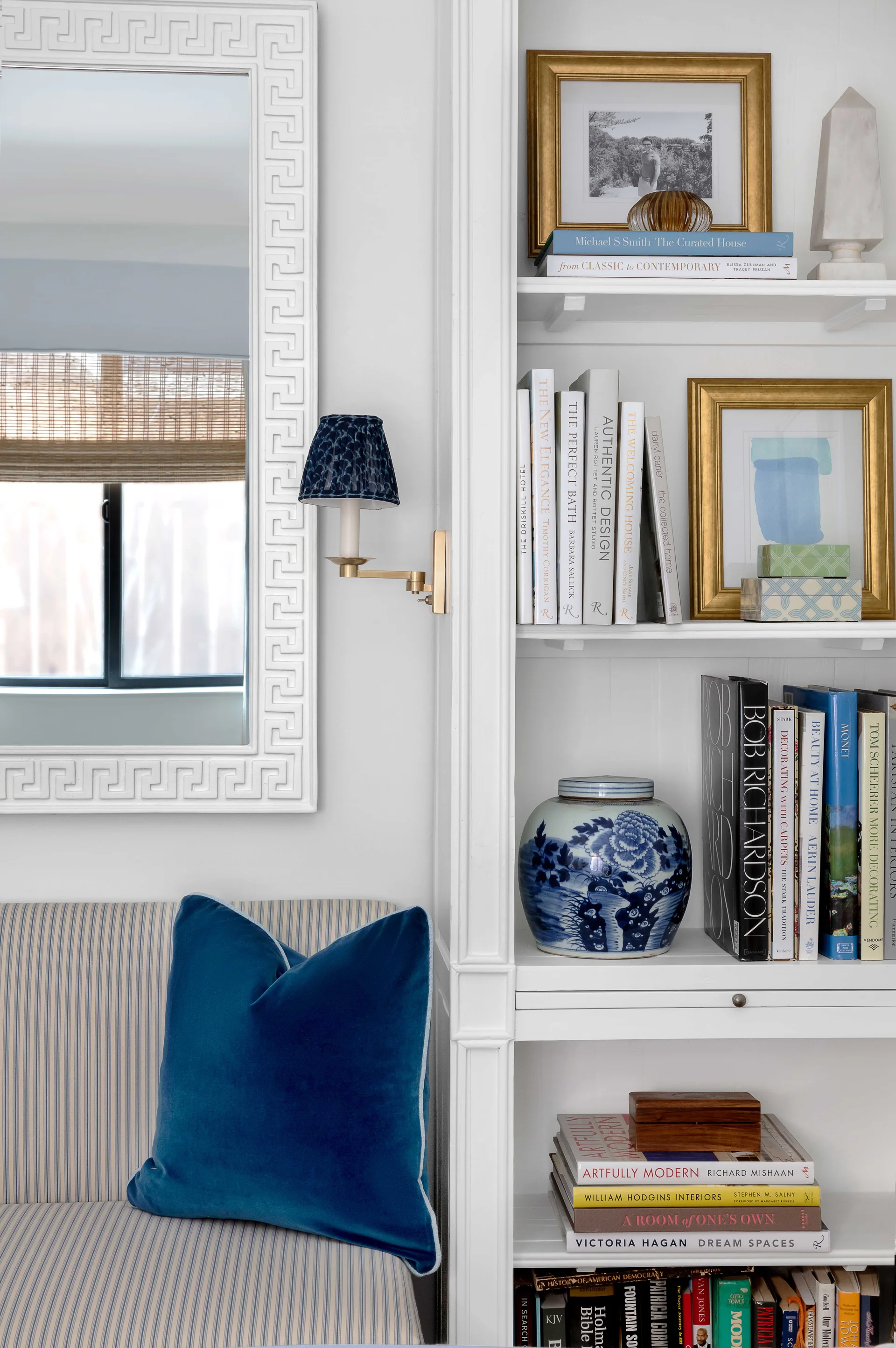 A white built-in bookshelf with books and a blue ginger jar sits beside a blue striped sofa and a Greek key patterned mirror.