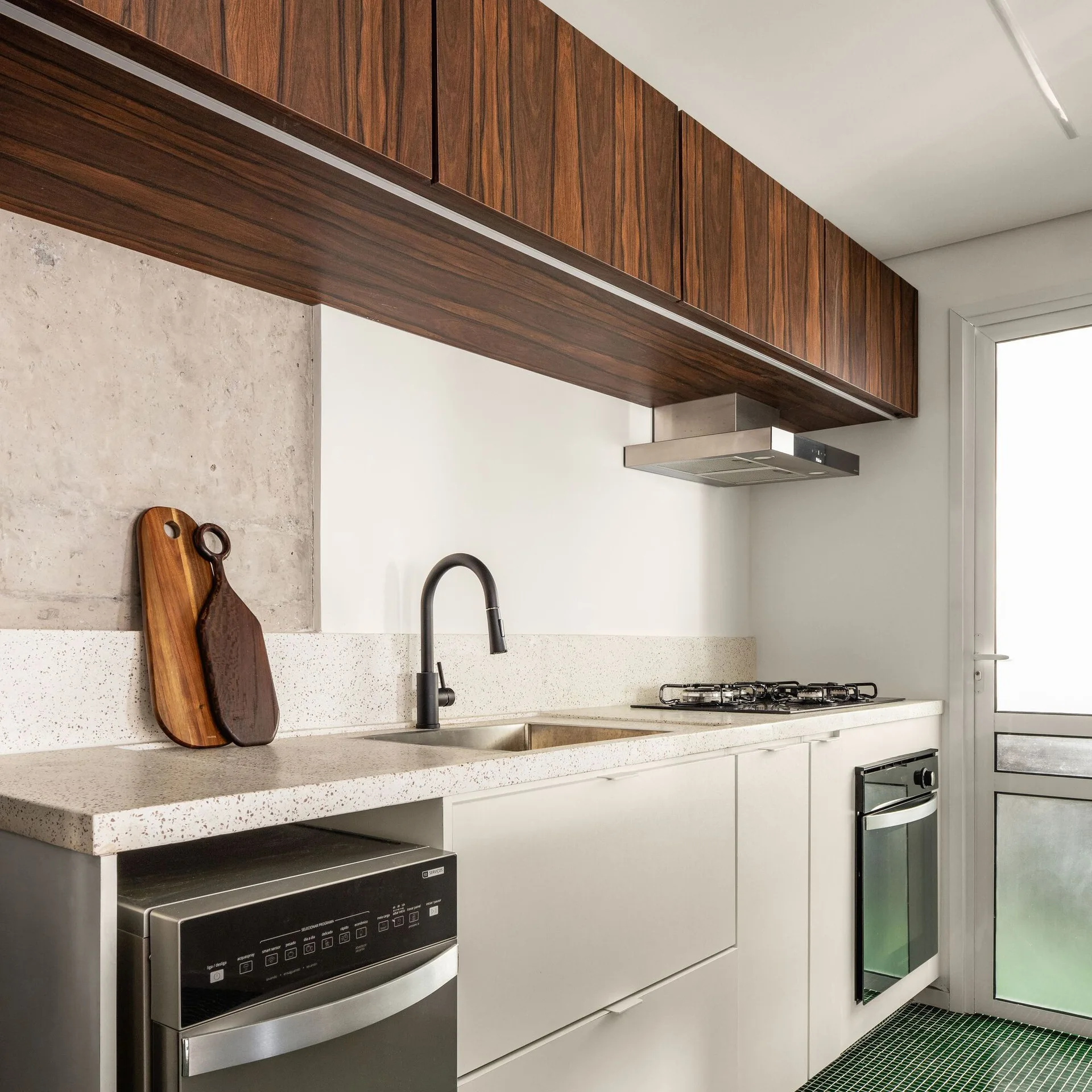 A modern kitchen featuring wood grain upper cabinets, white lower cabinets, a black faucet over a stainless steel sink, and green floor tiles.