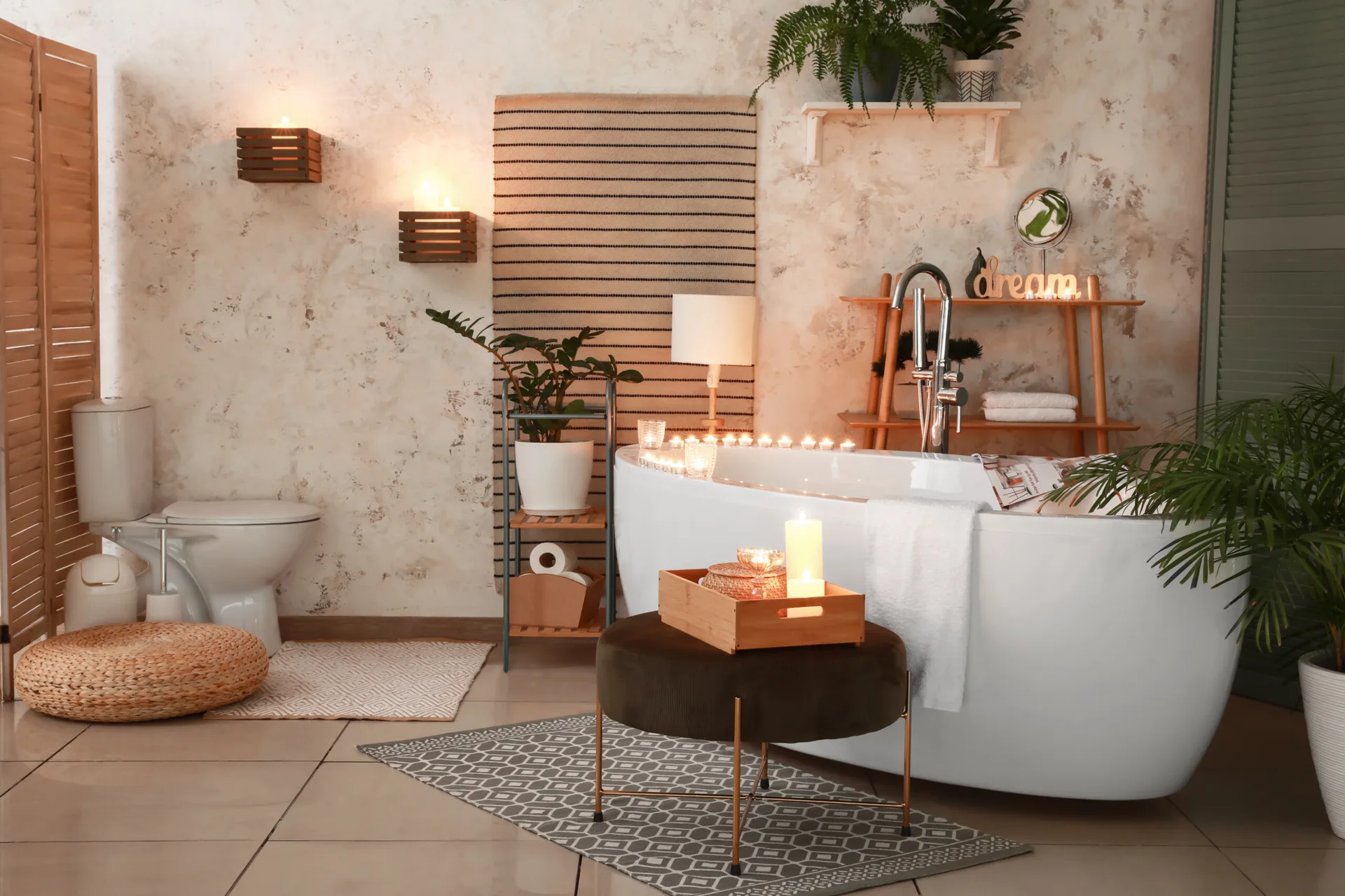 A candlelit bohemian bathroom features a white freestanding bathtub, textured beige walls, potted green plants, and a dark brown round ottoman.