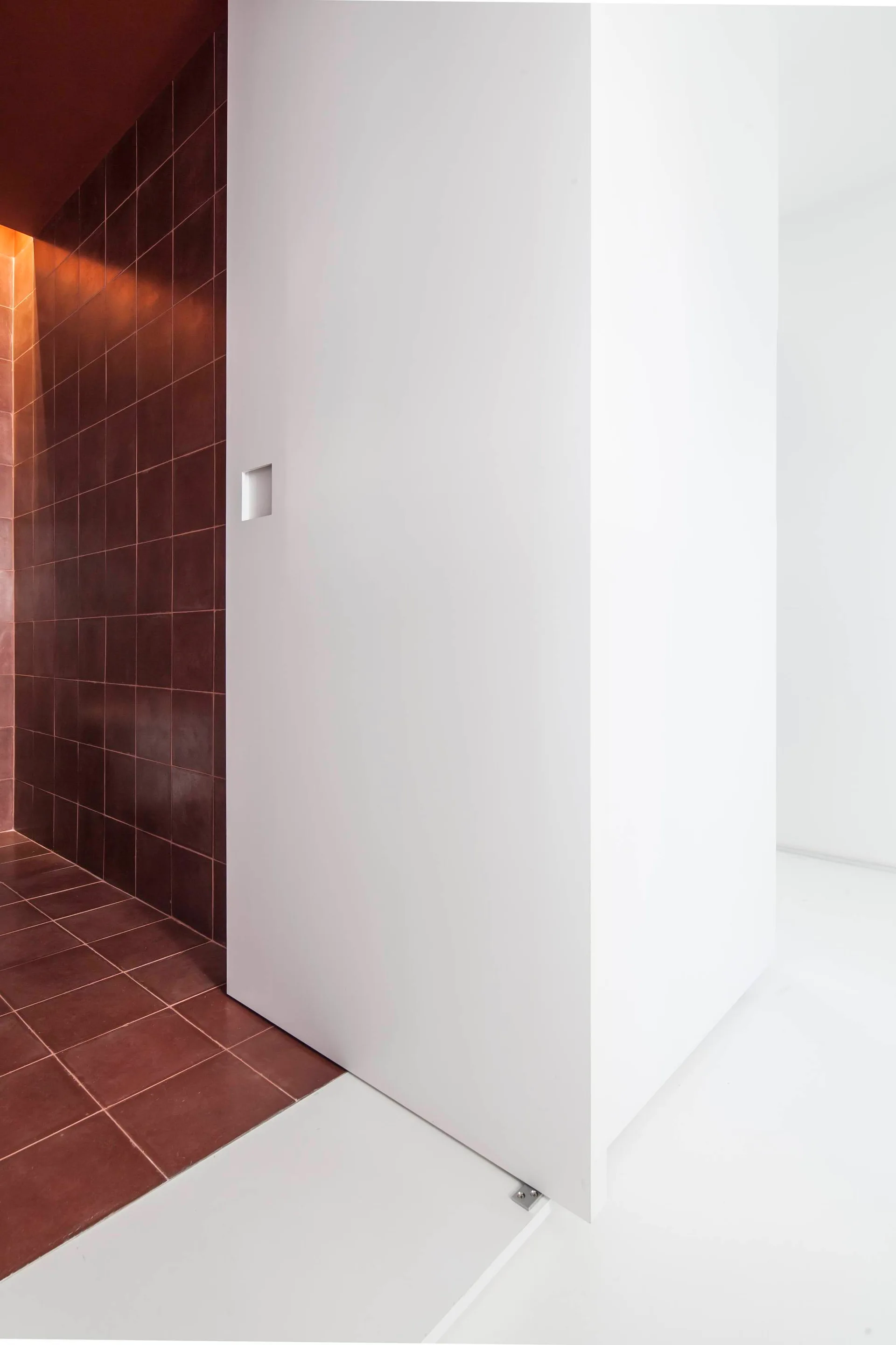 A white sliding door with a square recessed handle stands next to a dark red tiled wall and floor in a minimalist modern corridor.