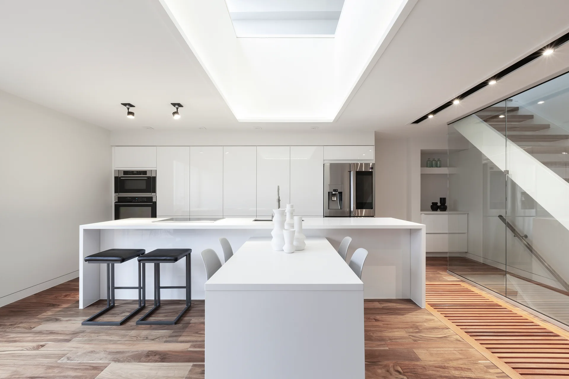 A minimalist white kitchen features glossy cabinetry, an island with seating, a large ceiling skylight, and a glass-walled staircase on wooden floors.