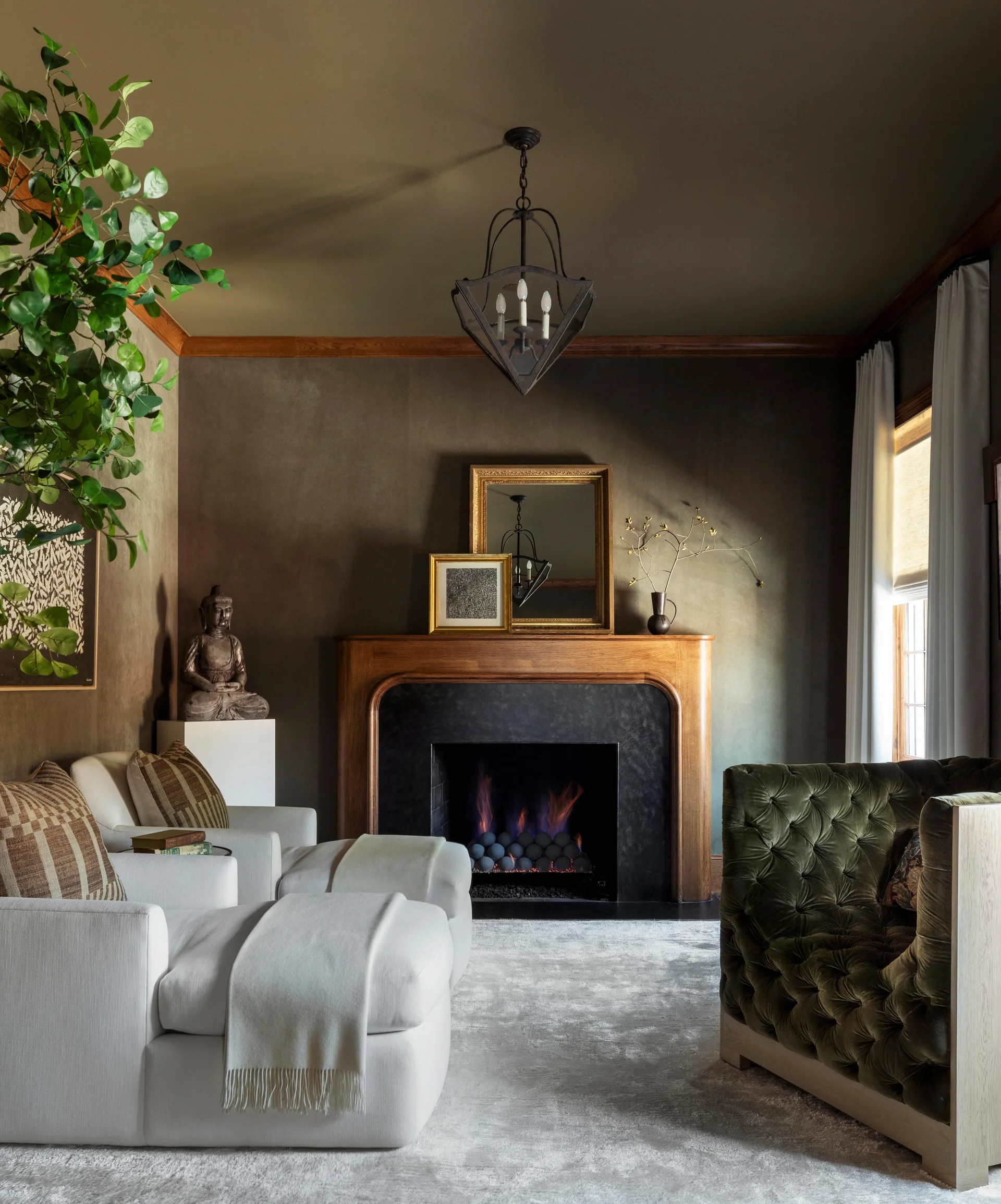 A dark olive green living room features two white armchairs with ottomans, a tufted green velvet sofa, and a wood fireplace mantel with a mirror.