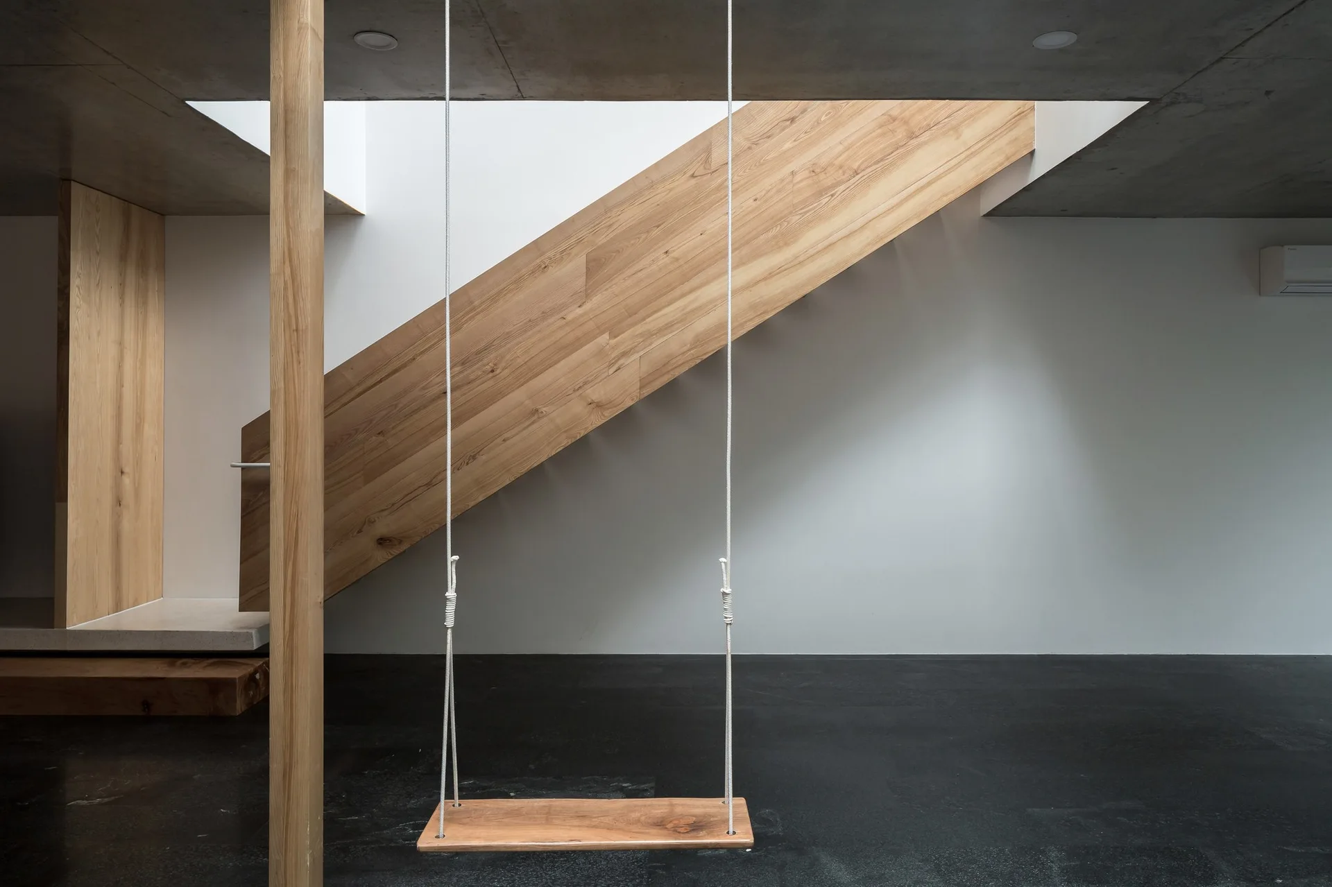 A minimalist interior space featuring a light wood staircase, a suspended wooden swing, a matching wooden column, a black floor, and a concrete ceiling.