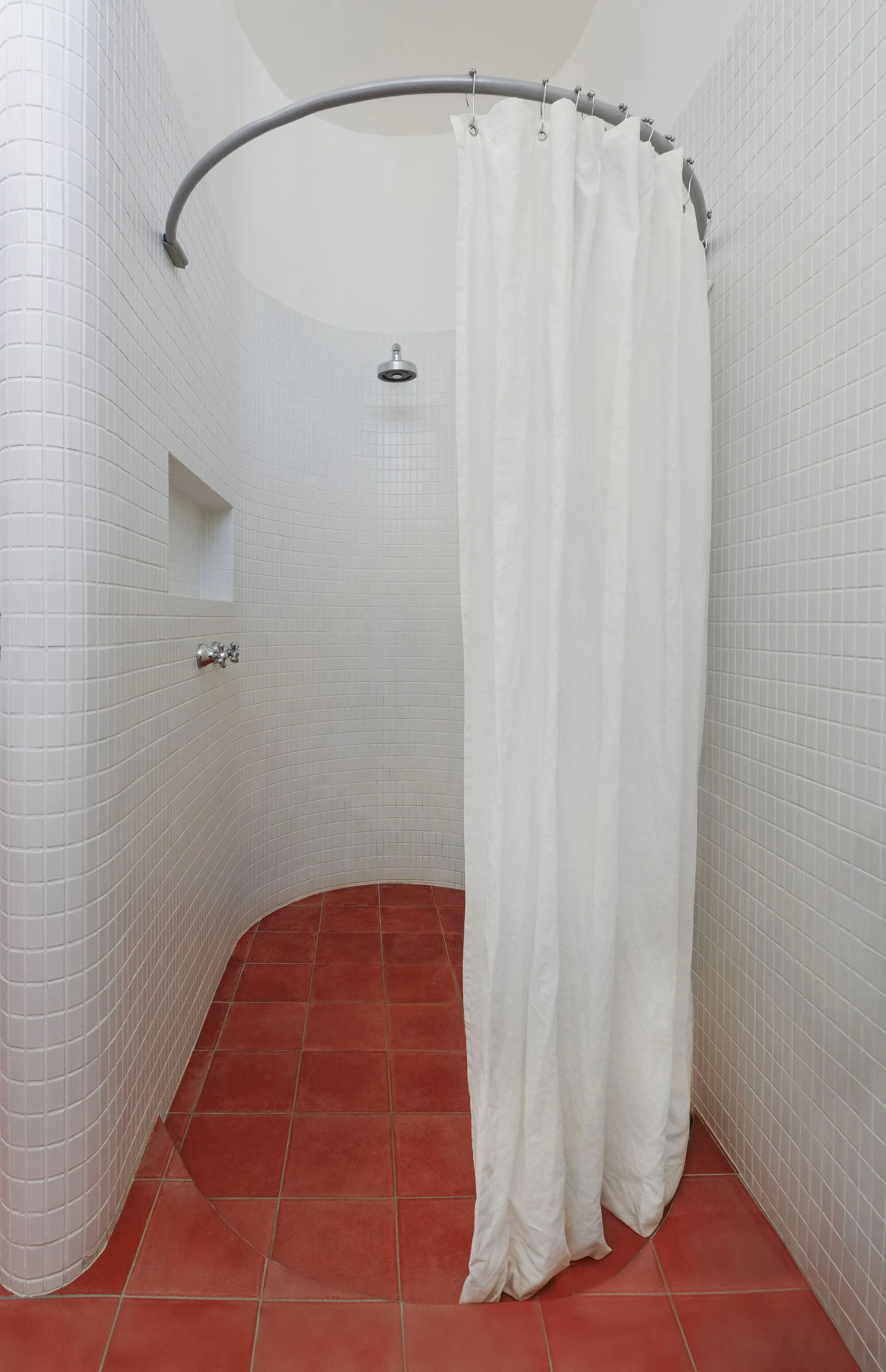 A curved walk-in shower features white square-tiled walls, a matching curved curtain rod, a white shower curtain, and red tiled flooring.