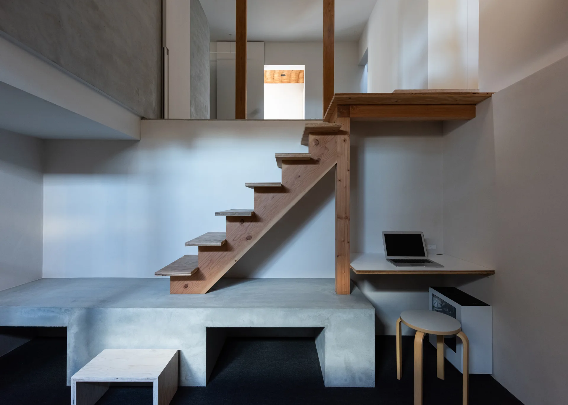 A wooden staircase rises from a grey concrete platform next to a minimalist built-in desk with a stool and laptop.
