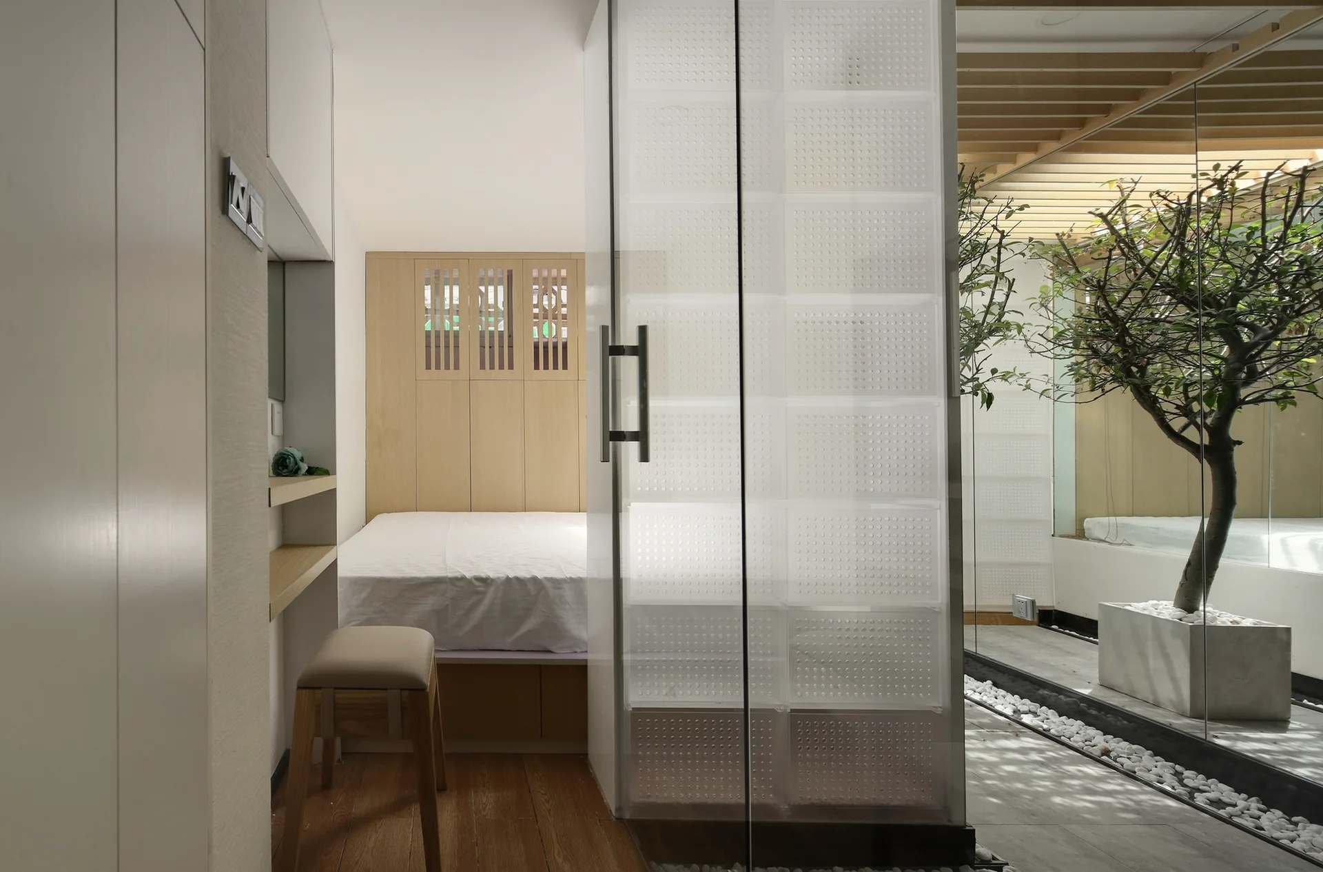 A minimalist bedroom featuring a platform bed and a translucent divider next to an indoor courtyard with a small tree and white pebbles.