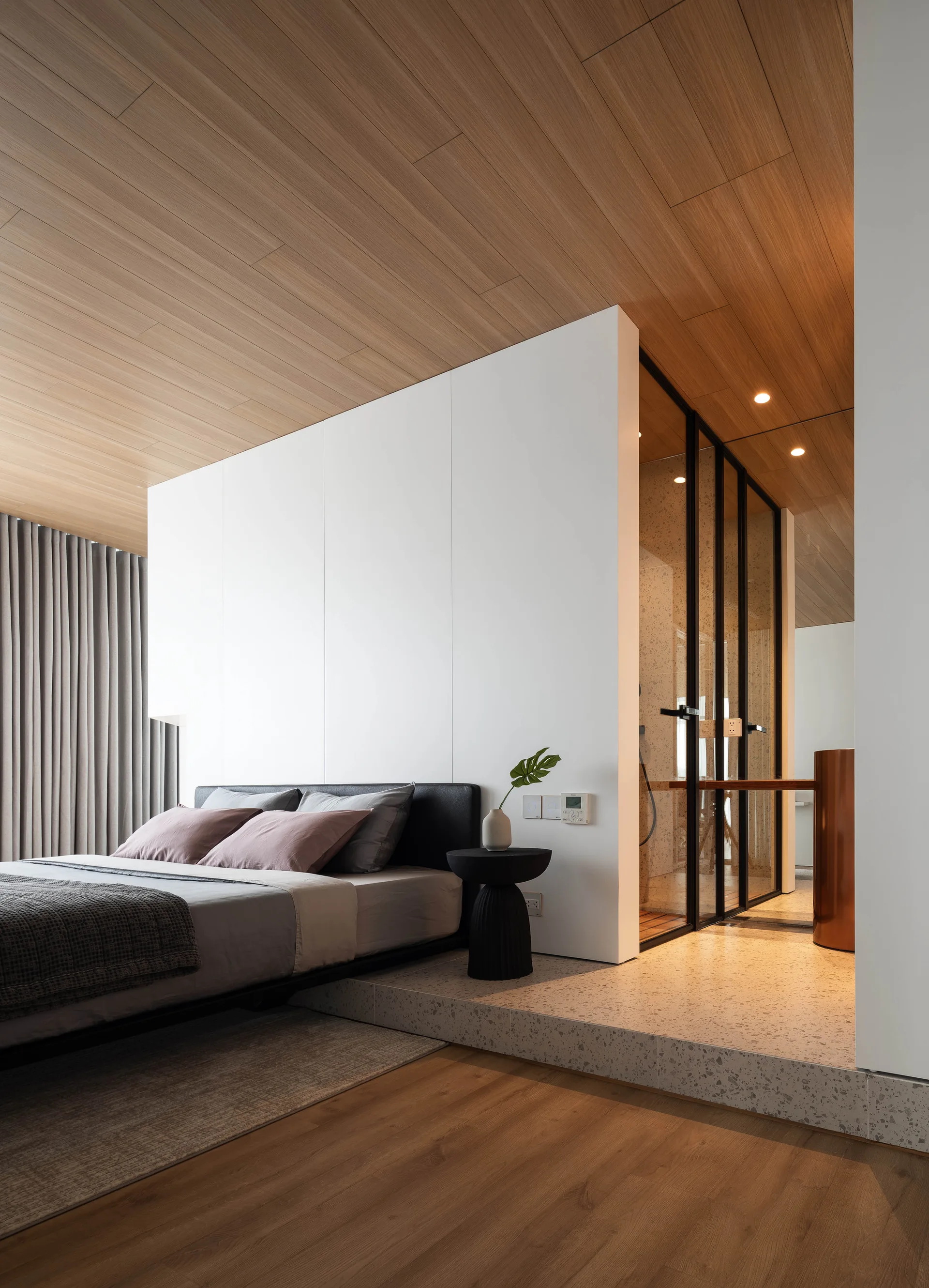 A modern bedroom features a wood plank ceiling, a low bed with gray pillows, and a raised terrazzo platform leading to a glass-enclosed bathroom.