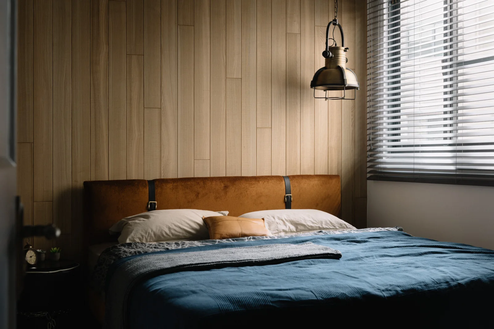 A bedroom features a brown velvet headboard against a vertical wood paneled wall with a blue bedspread and an industrial pendant light.