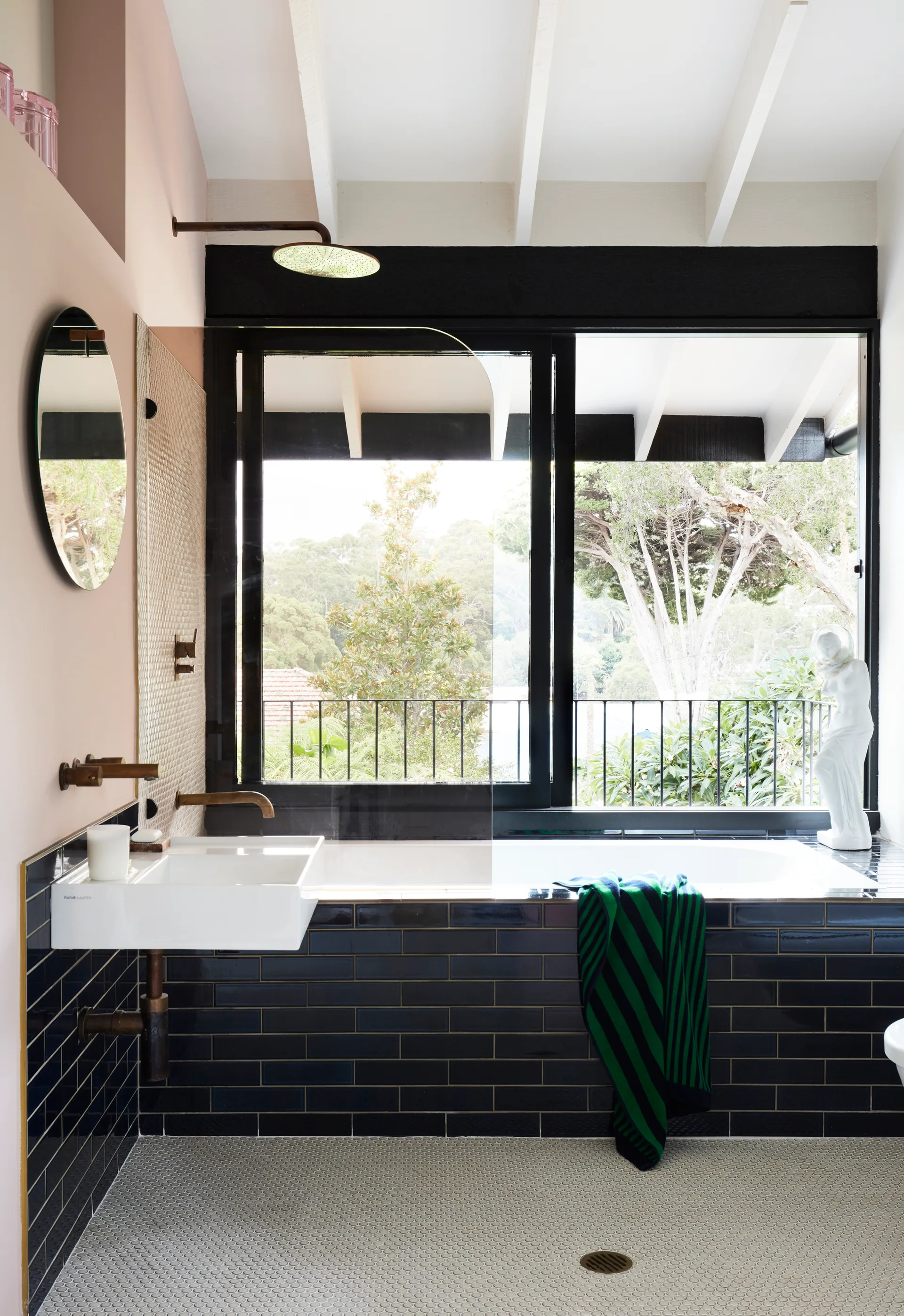 A modern bathroom features a bathtub enclosed in dark navy tiles, a wall-mounted sink with brass hardware, and a large window facing green trees.