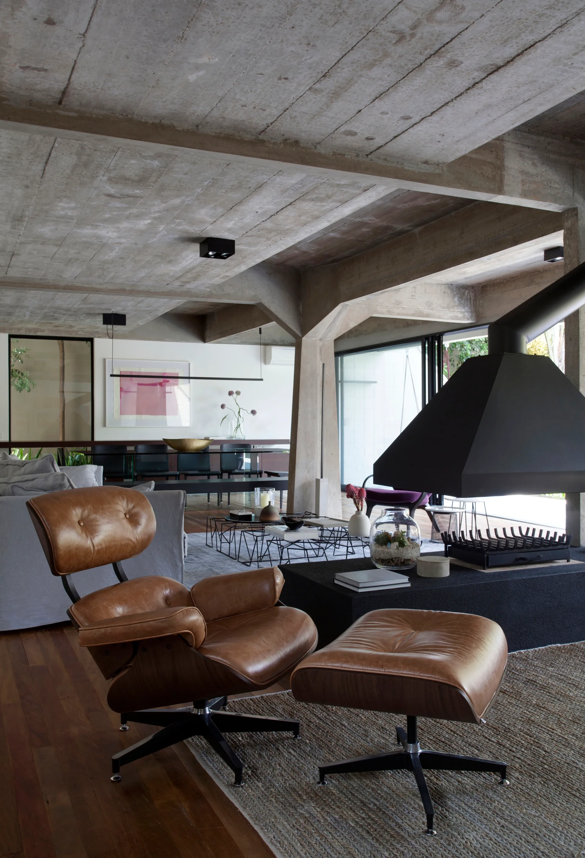 A modern living room features a brown leather lounge chair with ottoman, an exposed concrete ceiling, a black fireplace, and warm wood flooring.