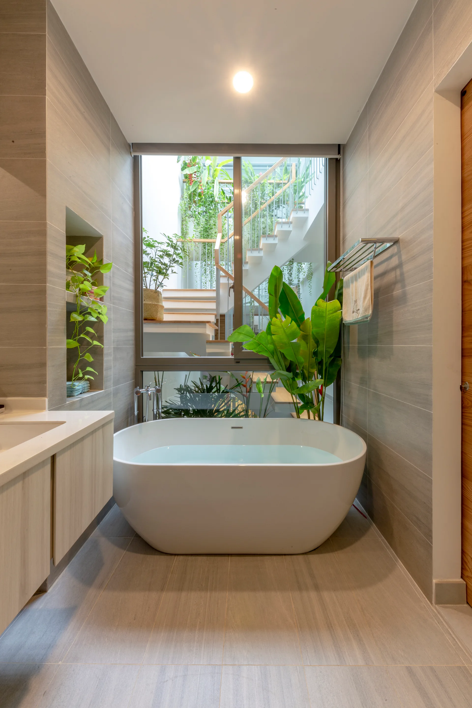 A modern bathroom features a white freestanding bathtub positioned in front of a large window overlooking a lush green courtyard with an open staircase.