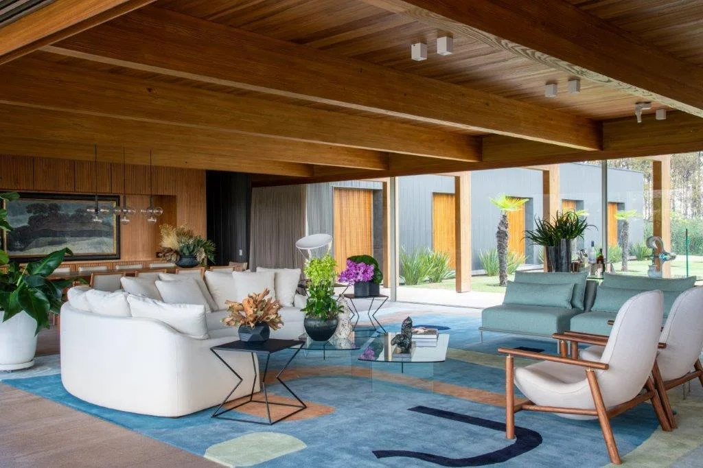 A modern open-plan living room features a curved white sofa, a large patterned blue rug, and a warm wood plank ceiling with exposed beams.