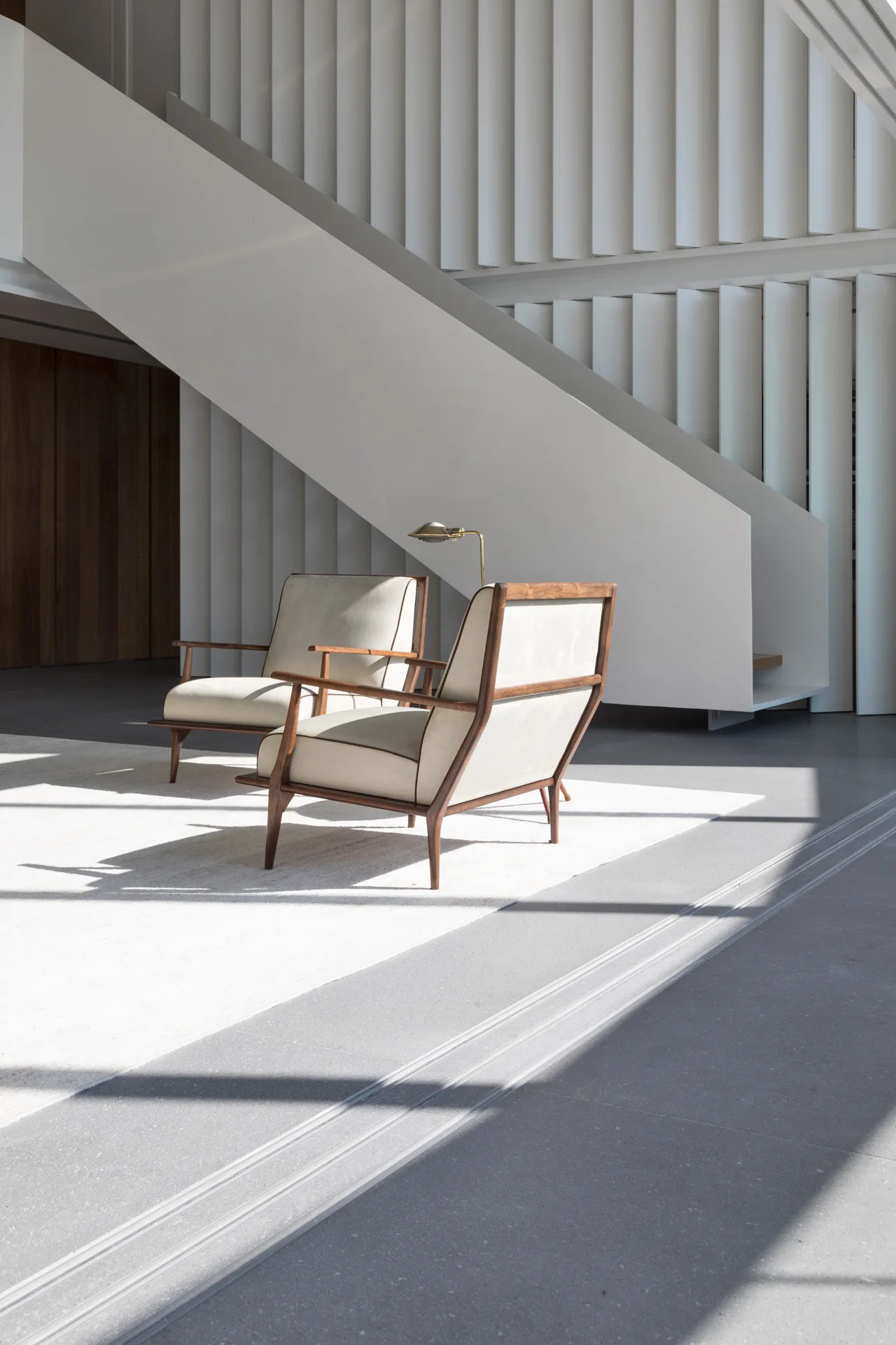 Two cream lounge chairs with wood frames sit on a light rug in front of a white geometric staircase and a vertical slatted wall.