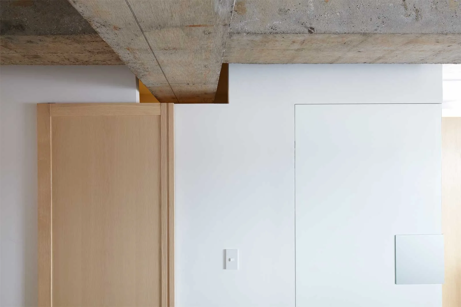 An architectural interior detail showing an exposed concrete ceiling beam above a smooth white plaster wall with a light wood door.