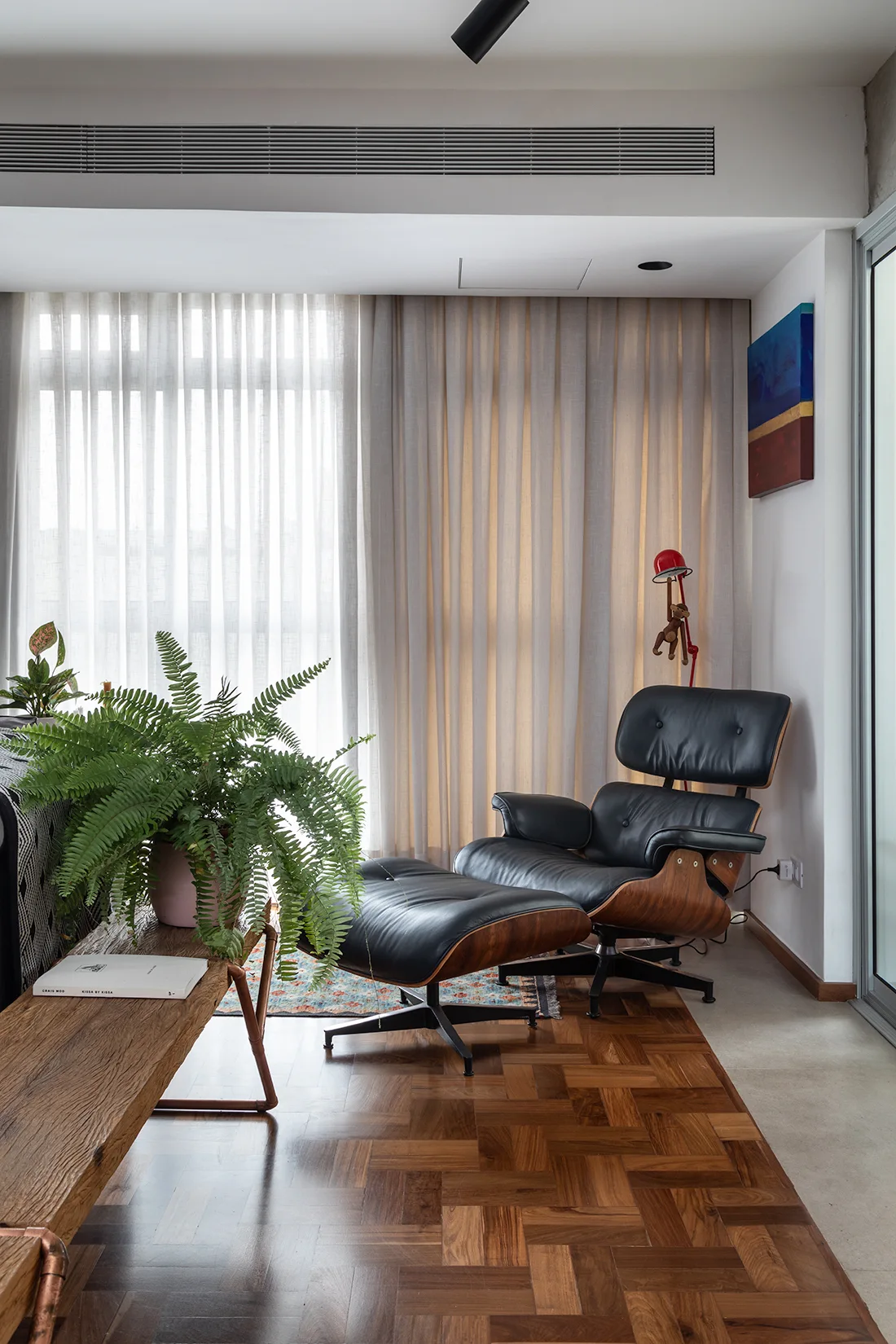 A black leather lounge chair and ottoman rest on herringbone wood floors next to sheer white curtains and a rustic wooden bench with a lush green fern.