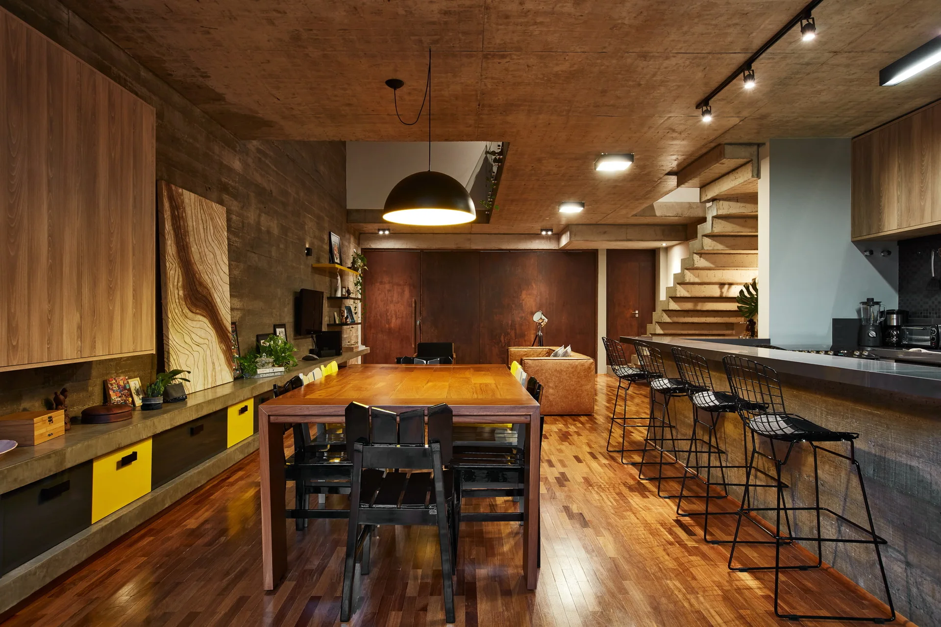 An open-plan dining space features a large wooden table with black chairs beneath a raw concrete ceiling and a kitchen island with wire bar stools.