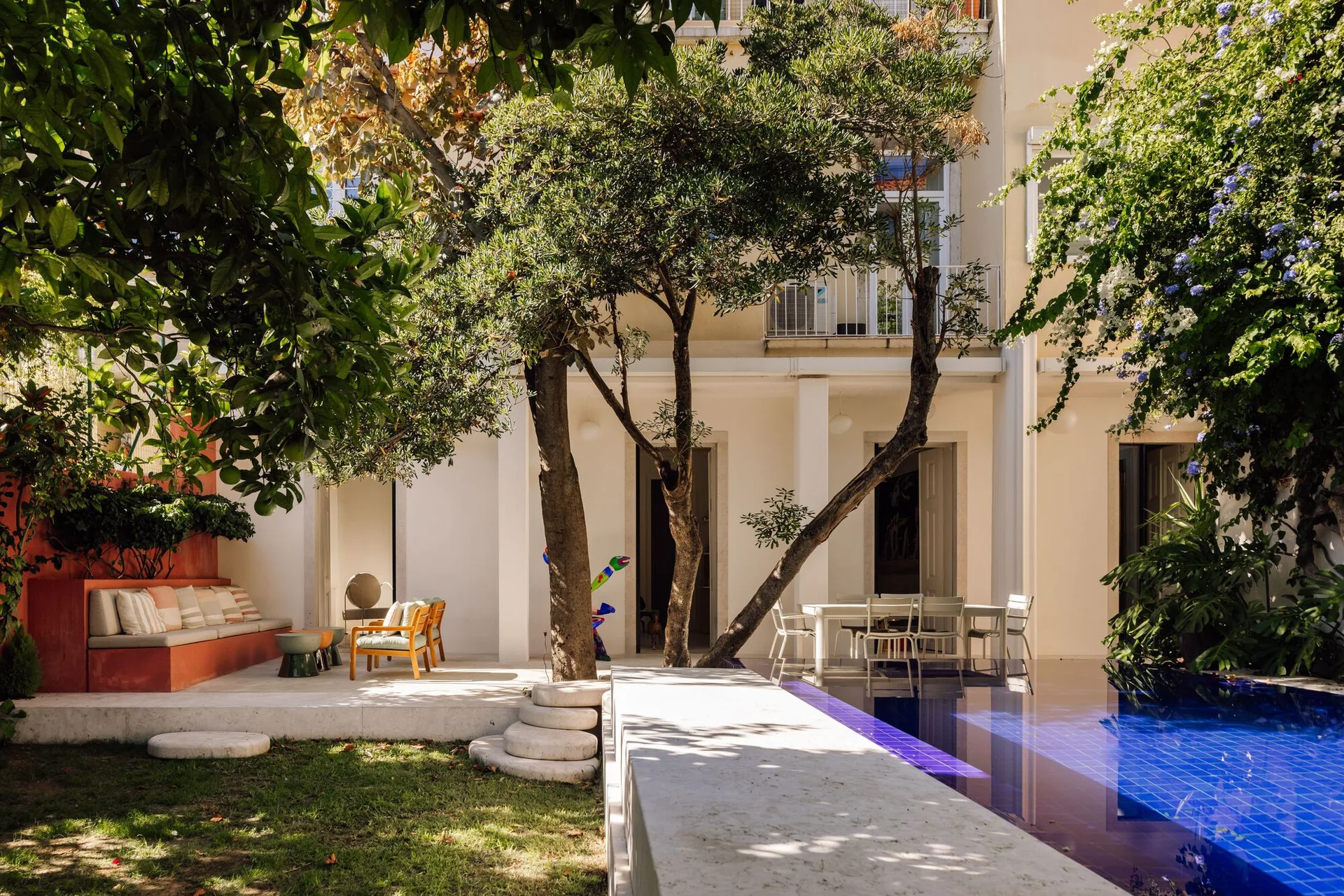 A sunny Mediterranean courtyard with a blue tiled swimming pool, a built-in terracotta plaster bench, and mature trees shading a paved platform.