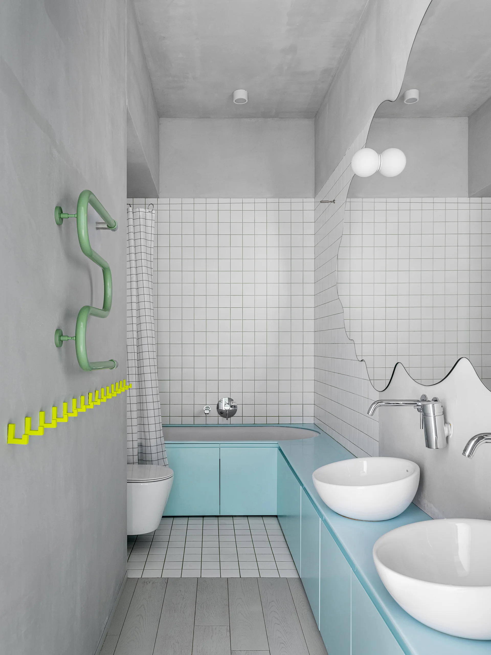A modern bathroom featuring a light blue vanity cabinet, a wavy wall mirror, white grid-pattern tiled walls, and bright yellow hooks.