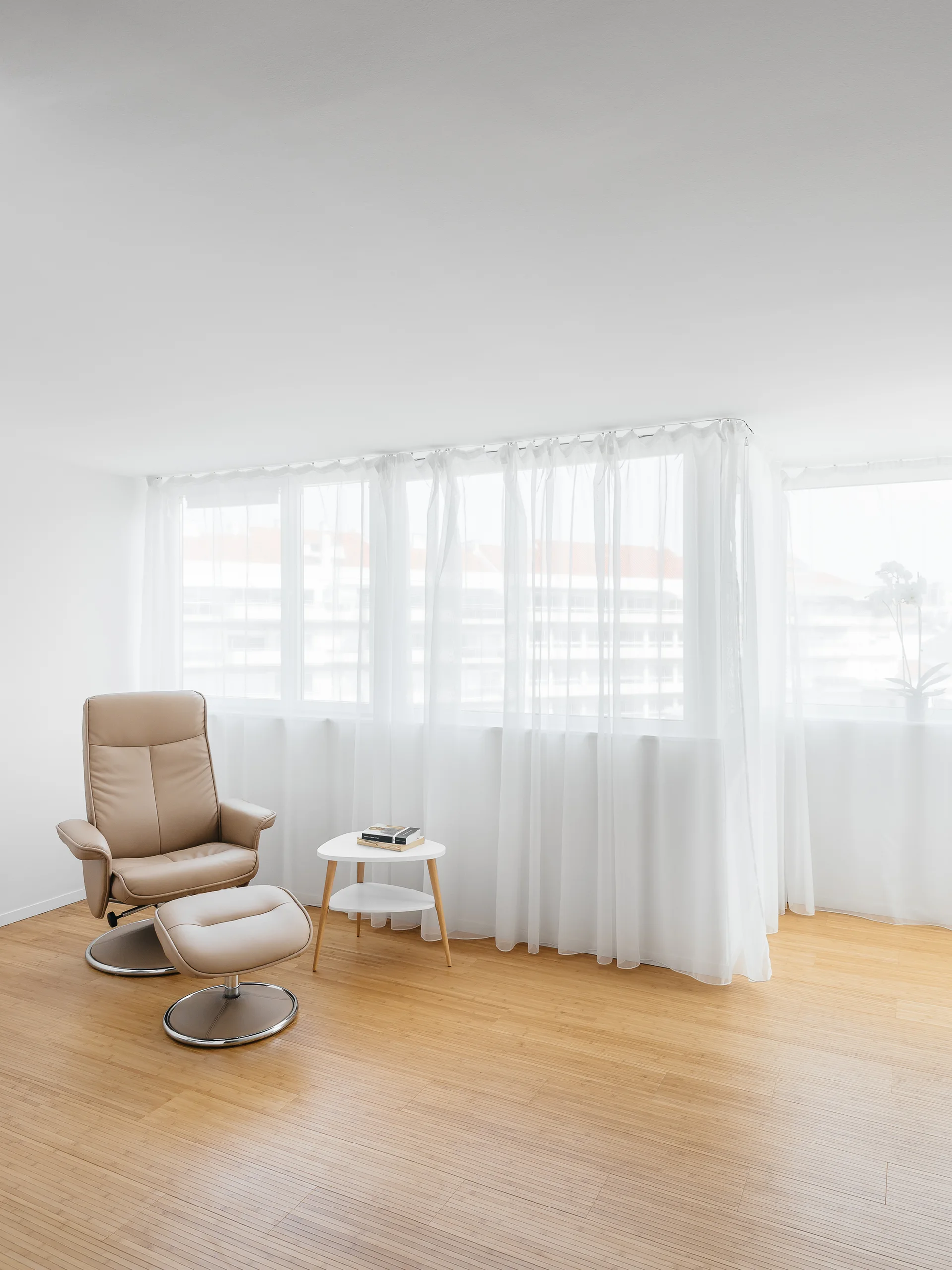 A bright minimalist room features a beige leather recliner and ottoman next to a white side table in front of a window with sheer white curtains.